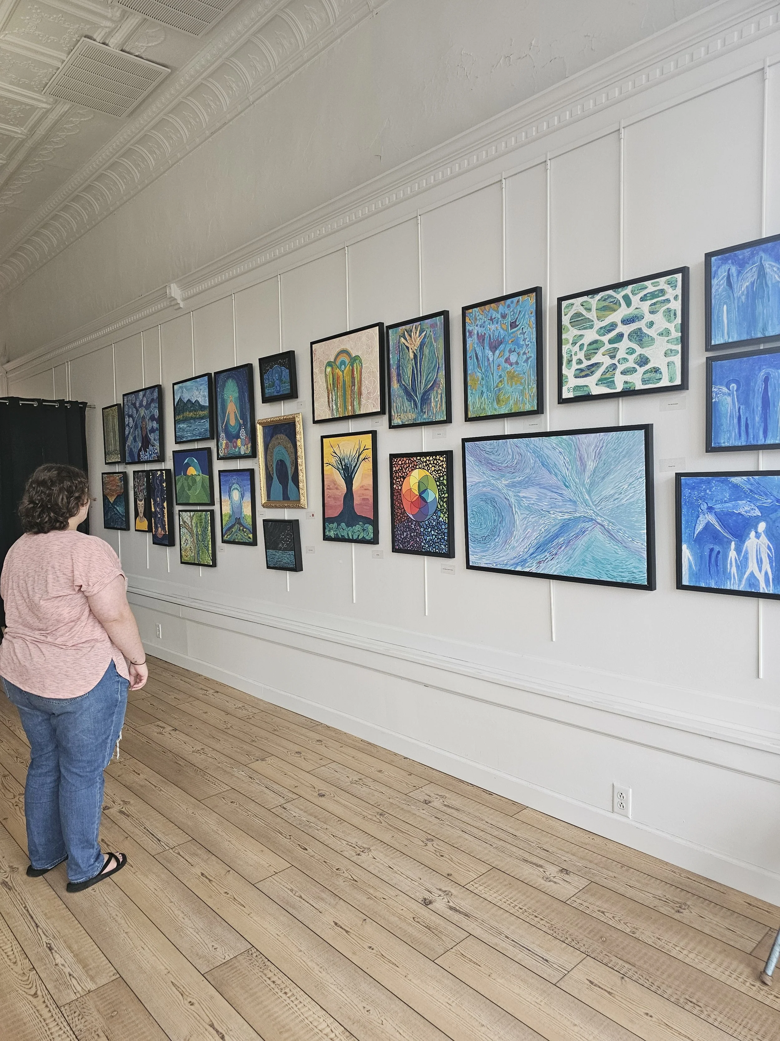 A woman viewing an art gallery wall filled with colorful paintings, including abstract and landscape styles, in a room with wooden floors and decorative ceiling molding.