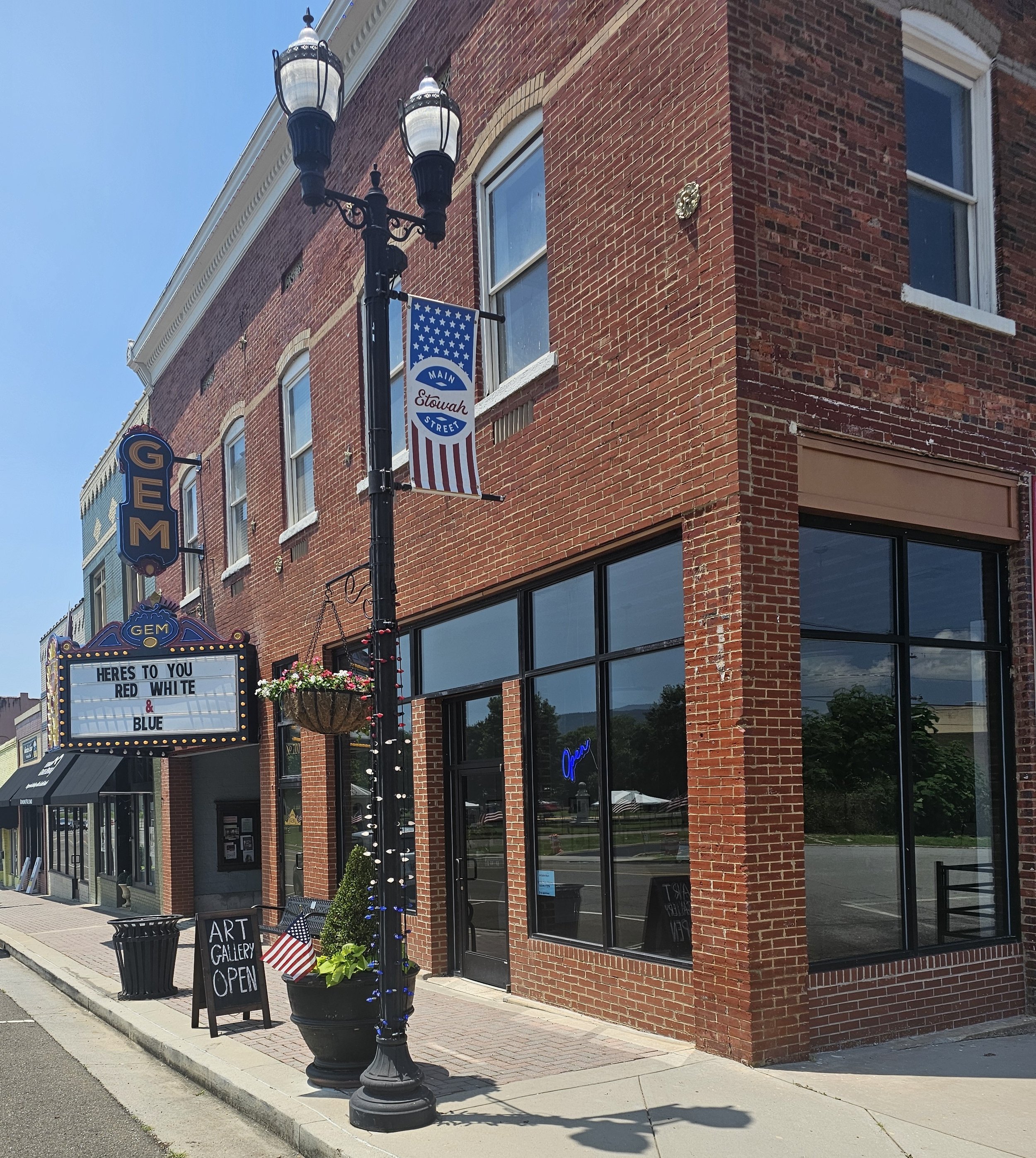Sidewalk view of a red brick art gallery building with a large window, a neon 'Open' sign, outdoor decor including a small American flag and a flower pot, a lit sign announcing 'Art Gallery Open', and a marquee sign that reads 'Here's to you, red, white, and blue'. A streetlamp with decorative lighting and banners, one American flag themed, are also visible.