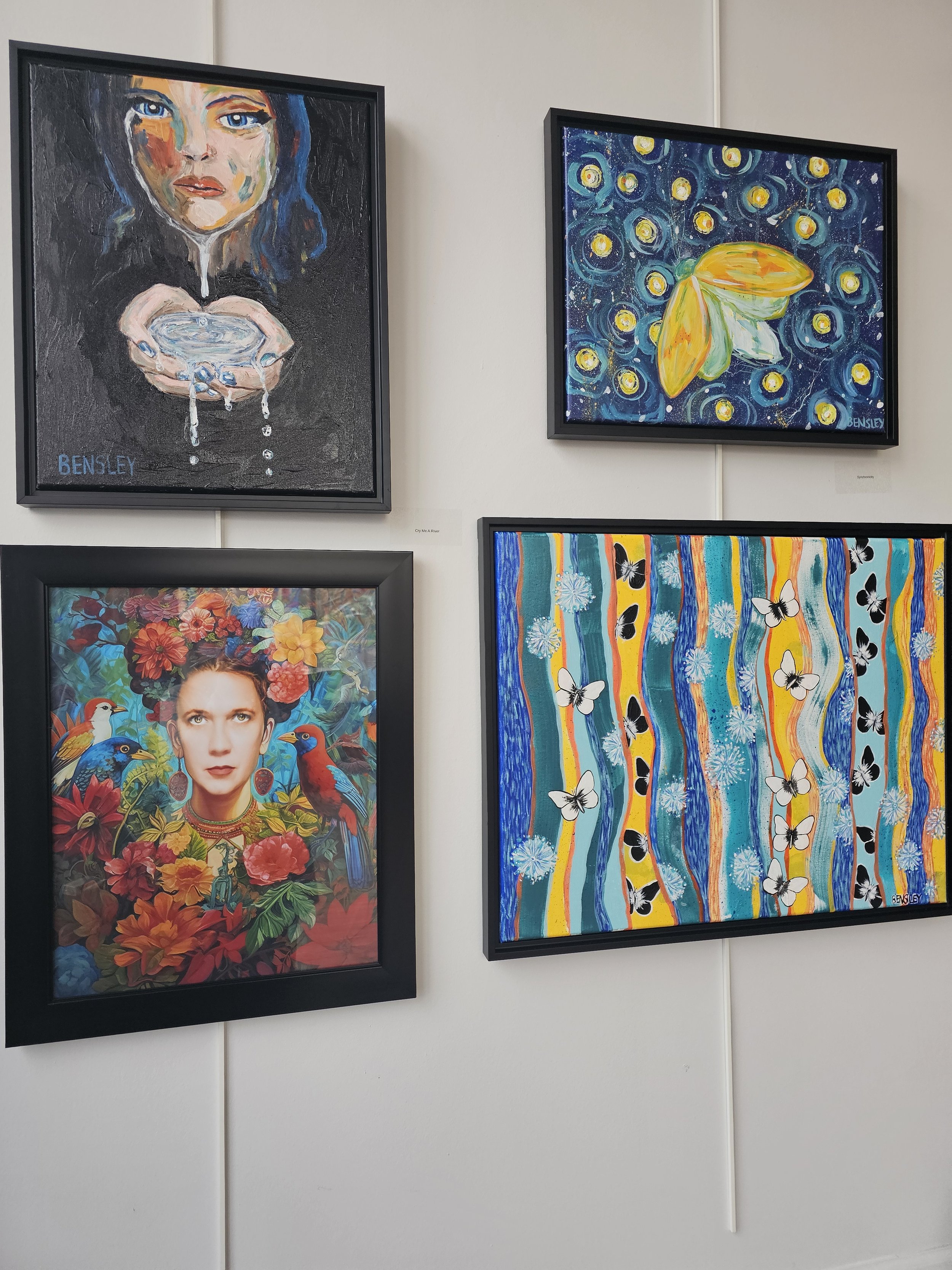 Four colorful paintings on a white wall, including a portrait of a woman surrounded by flowers and birds, a floral scene with tropical birds and flowers, an abstract butterfly against a starry background, and an abstract artwork with wavy lines and butterflies.