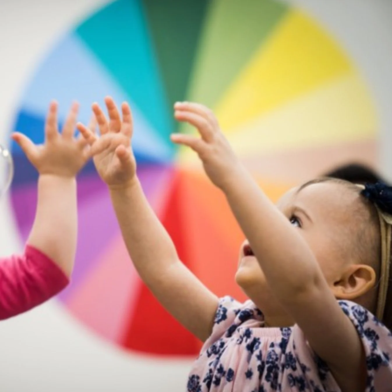A young girl reaching out her hands towards another child's hands with a colorful rainbow umbrella in the background.