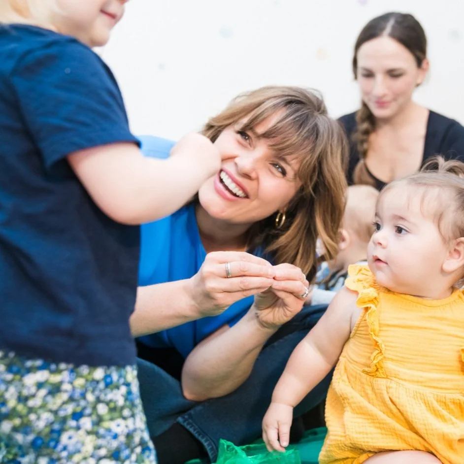 A woman plays with a young girl in a classroom or daycare setting, while another woman and children look on, engaging with each other.