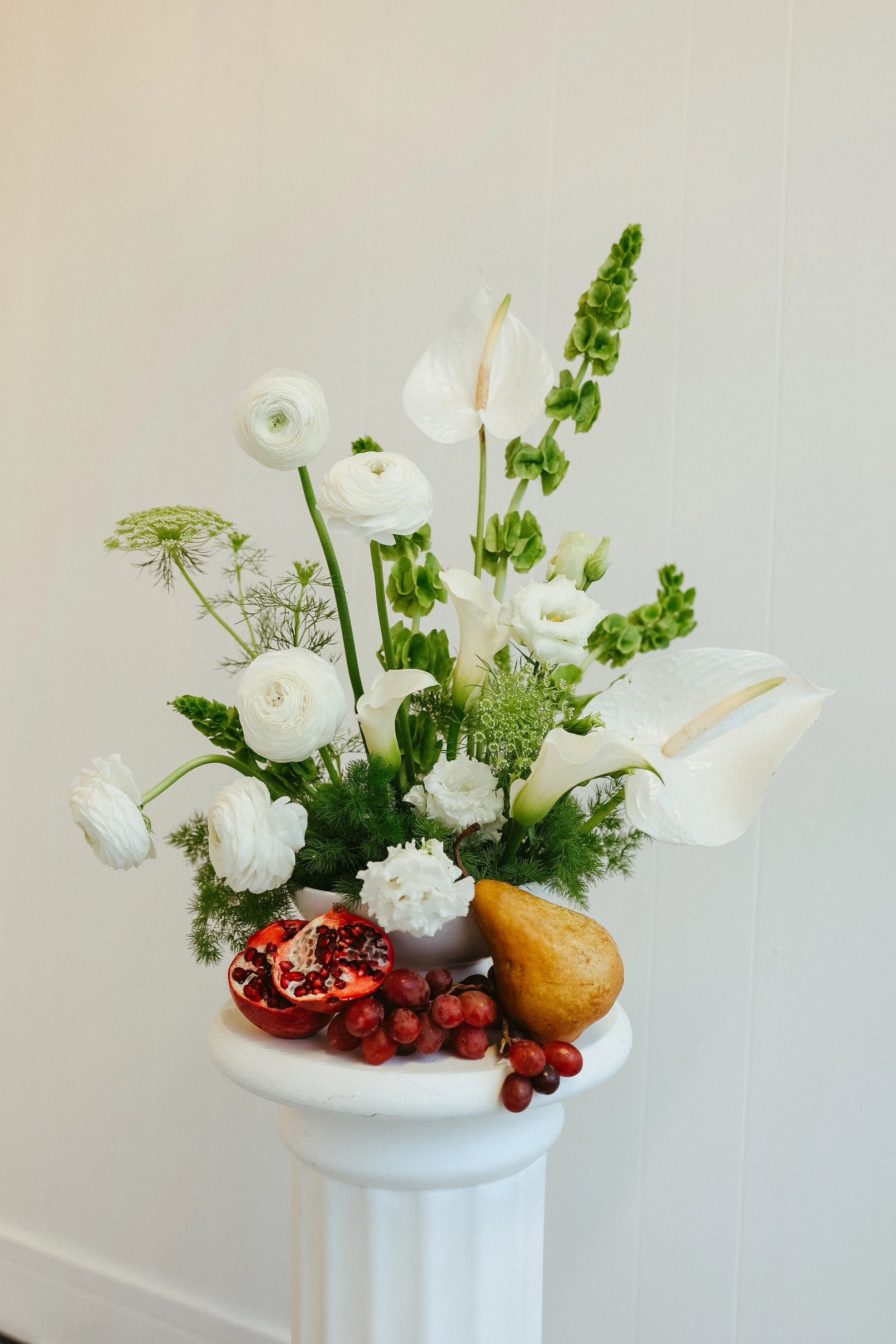 A white floral arrangement in a white vase with various white flowers and greenery, placed on a white pedestal with pomegranate, grapes, and a pear in front of it against a plain beige background.