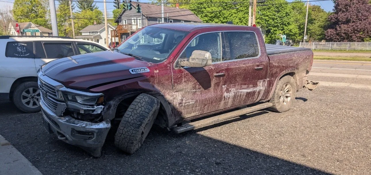 Red pickup truck with front-end damage and a flat front left tire, parked on the side of a street next to a white car, with houses and trees in the background.