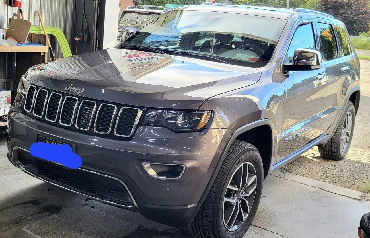 A gray Jeep Grand Cherokee SUV parked in a driveway next to a garage with tools and equipment visible on a workbench. The vehicle is clean and shiny, with a New York license plate.