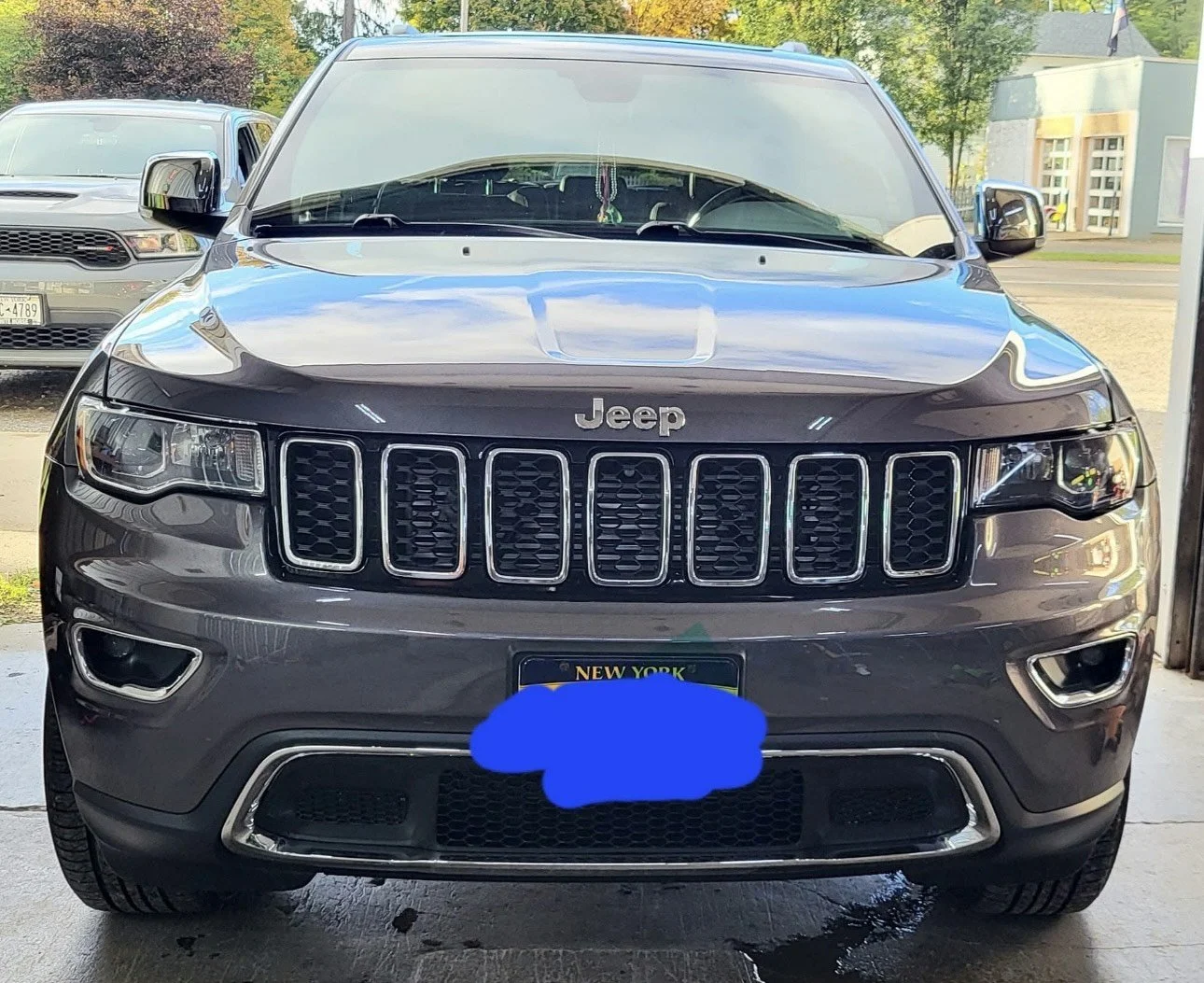 Front view of a gray Jeep SUV parked in a garage with another car and trees in the background.