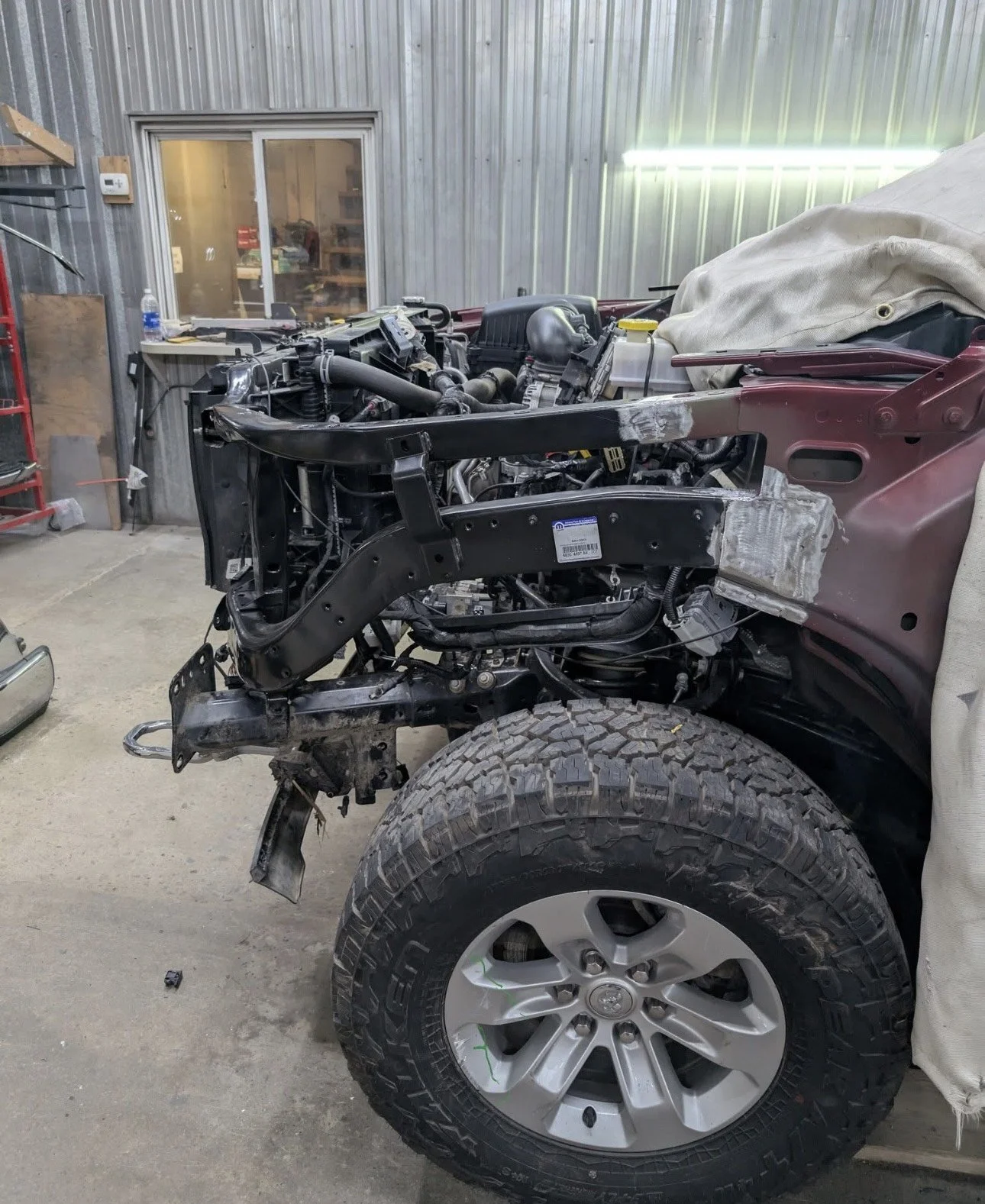 A partially assembled truck in a garage or workshop. The front part of the truck is missing its hood and other components, exposing the engine and chassis. The front wheel and tire are installed, but the area around the engine is unfinished with visi