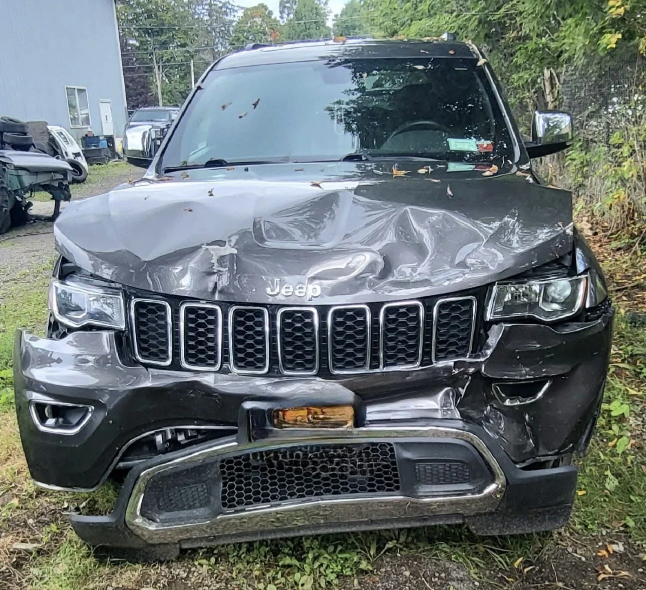 Front view of a black Jeep SUV with significant damage to the hood, grille, and front bumper after a crash, parked on grass near trees and other vehicles.