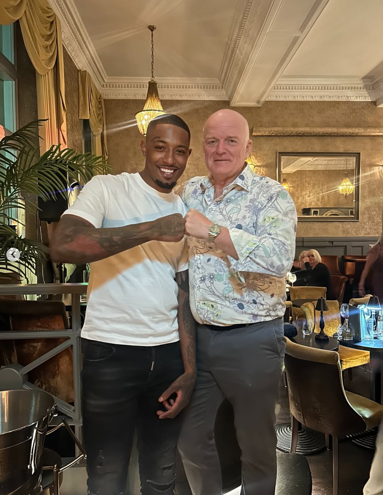 Two men are fist-bumping inside a decorated restaurant, smiling at the camera. One man is young with dark skin, tattoos, and a short haircut, while the other man is older, bald, and wearing a floral shirt. The background shows a mirror, warm lighting, and other diners.