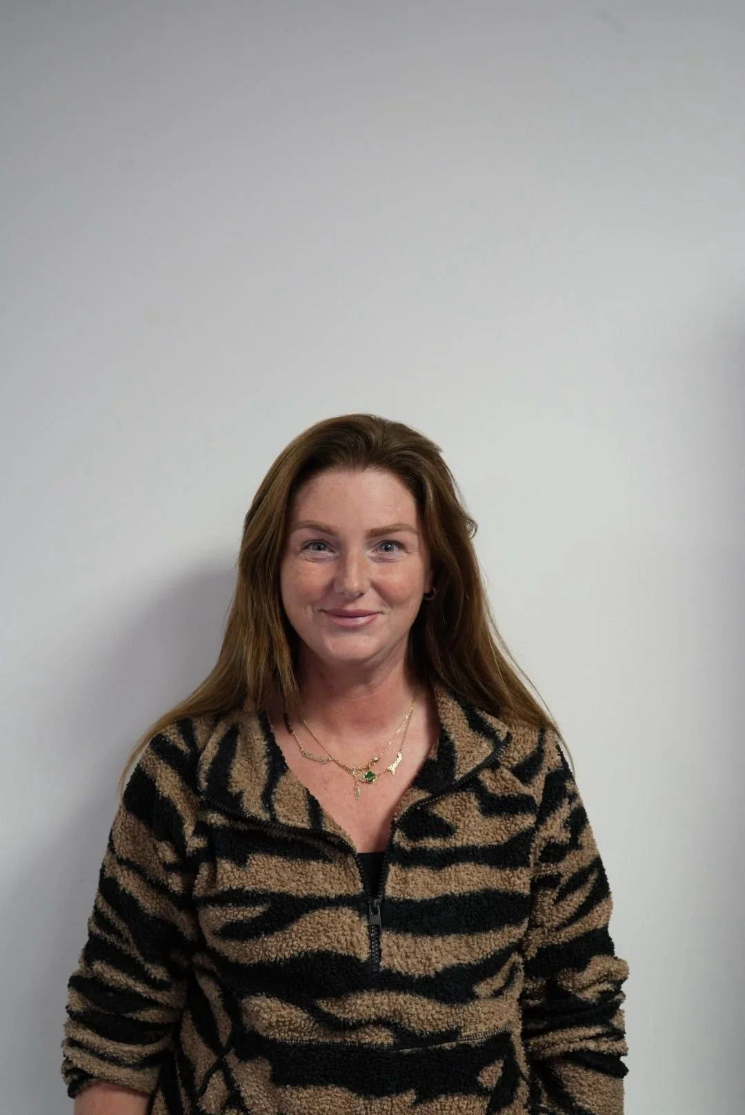 A woman with long brown hair wearing a black and tan tiger print fleece jacket, standing against a plain white wall.