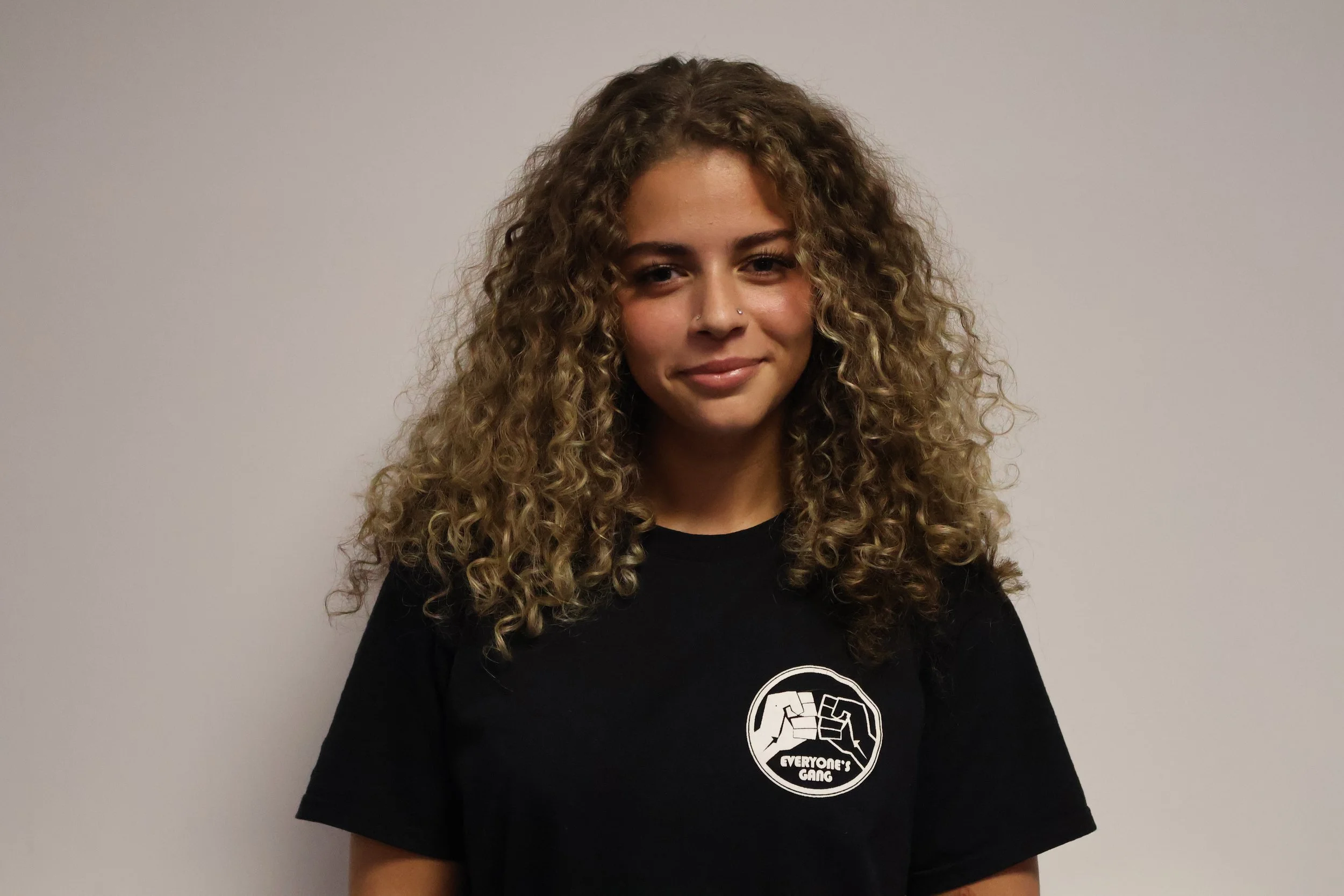 A young woman with curly black hair smiling and wearing glasses, hoop earrings, and layered necklaces in an indoor setting.