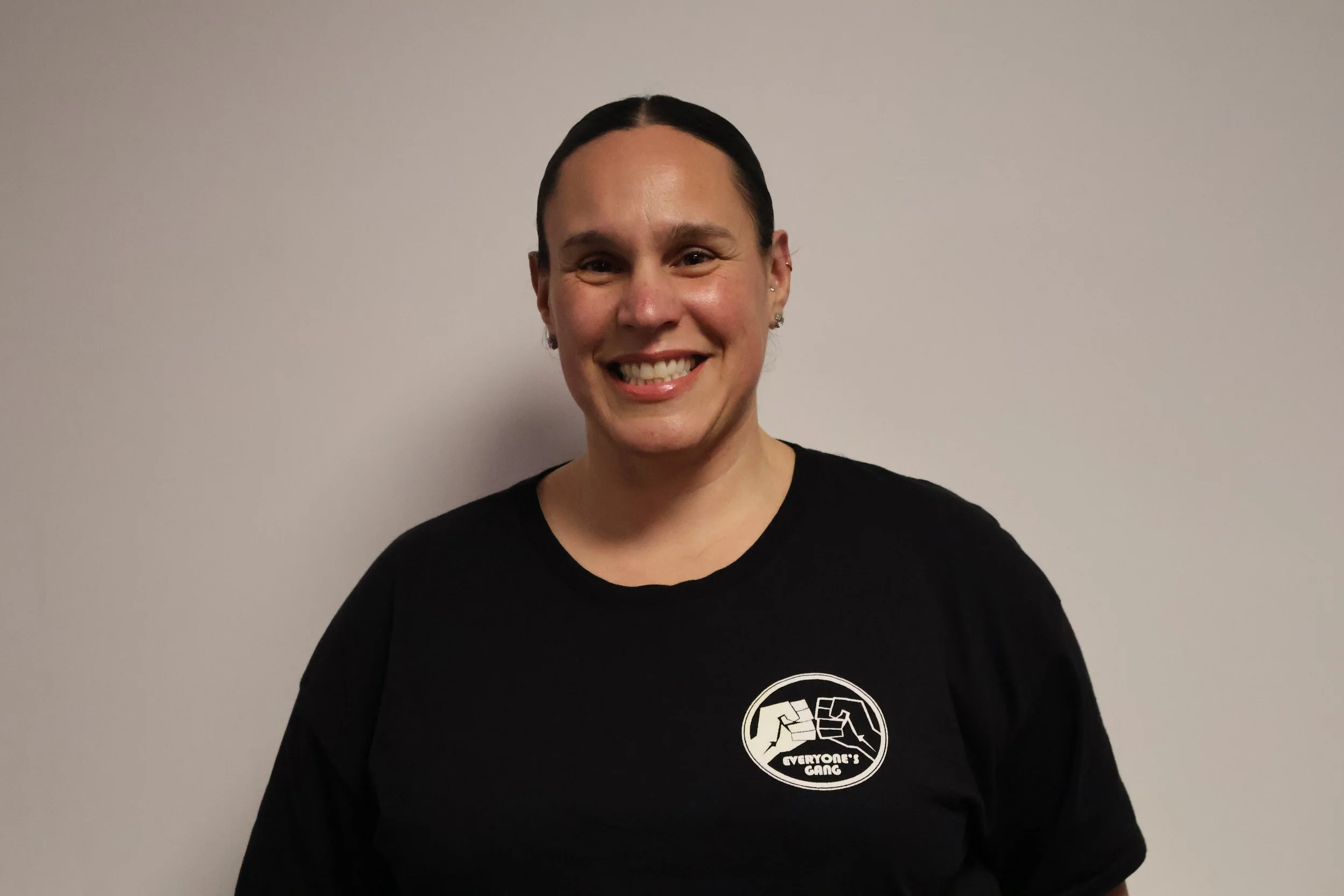 A man with glasses, facial hair, and dark wavy hair, sitting indoors with a neutral background and a plant in the distance.