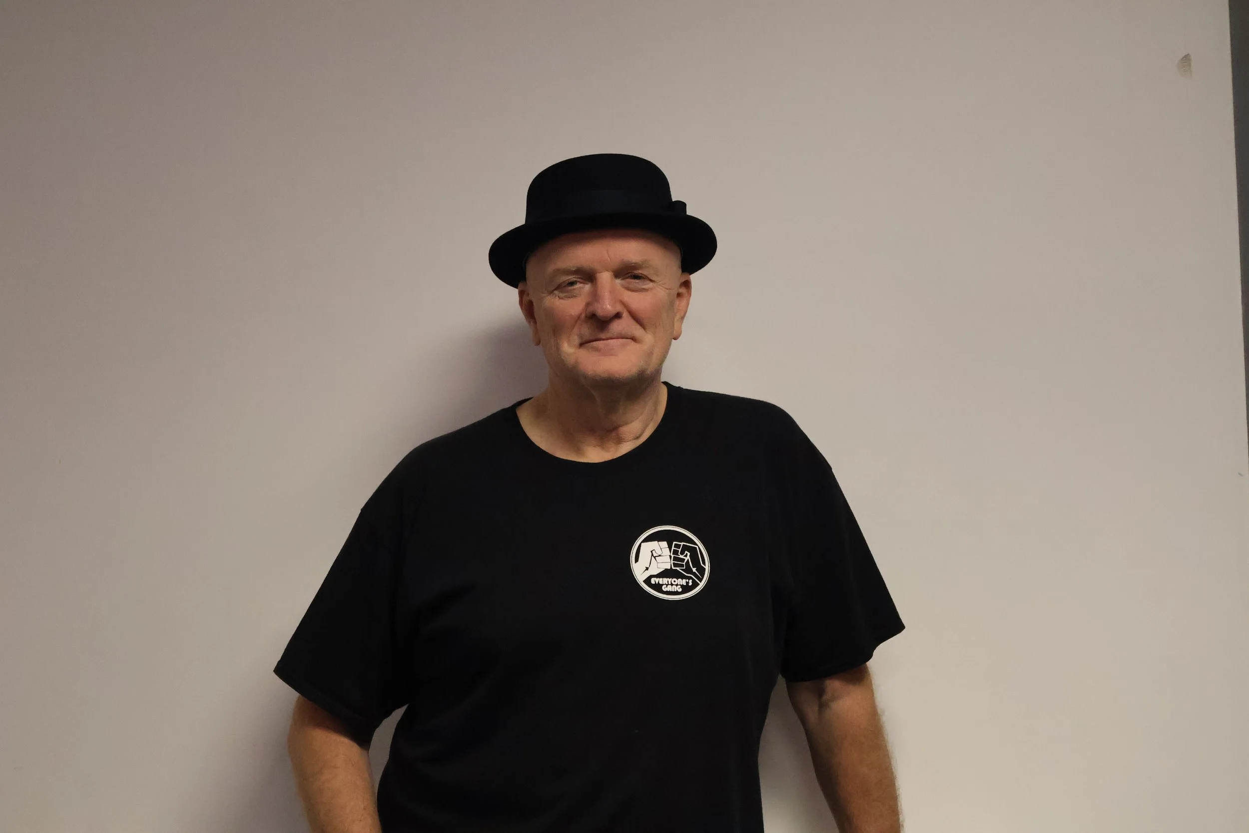 An man wearing a black hat and black t-shirt standing against a plain white wall.
