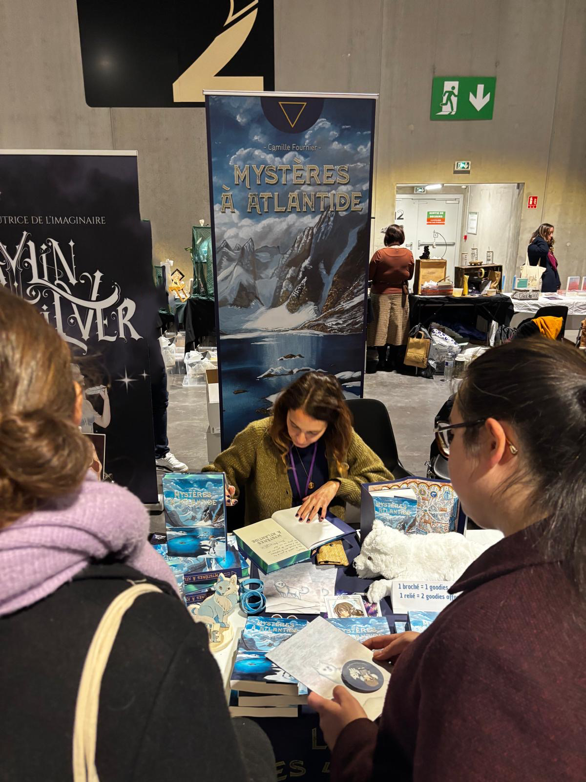 Une femme signe des livres à une table lors d'une séance de dédicaces pour le livre intitulé "Mystères à Atlantide". Deux autres personnes sont présentes, une avec un manteau violet et une autre avec une veste marron. En arrière-plan, un panneau promotionnel représentant un paysage montagneux et un lac.