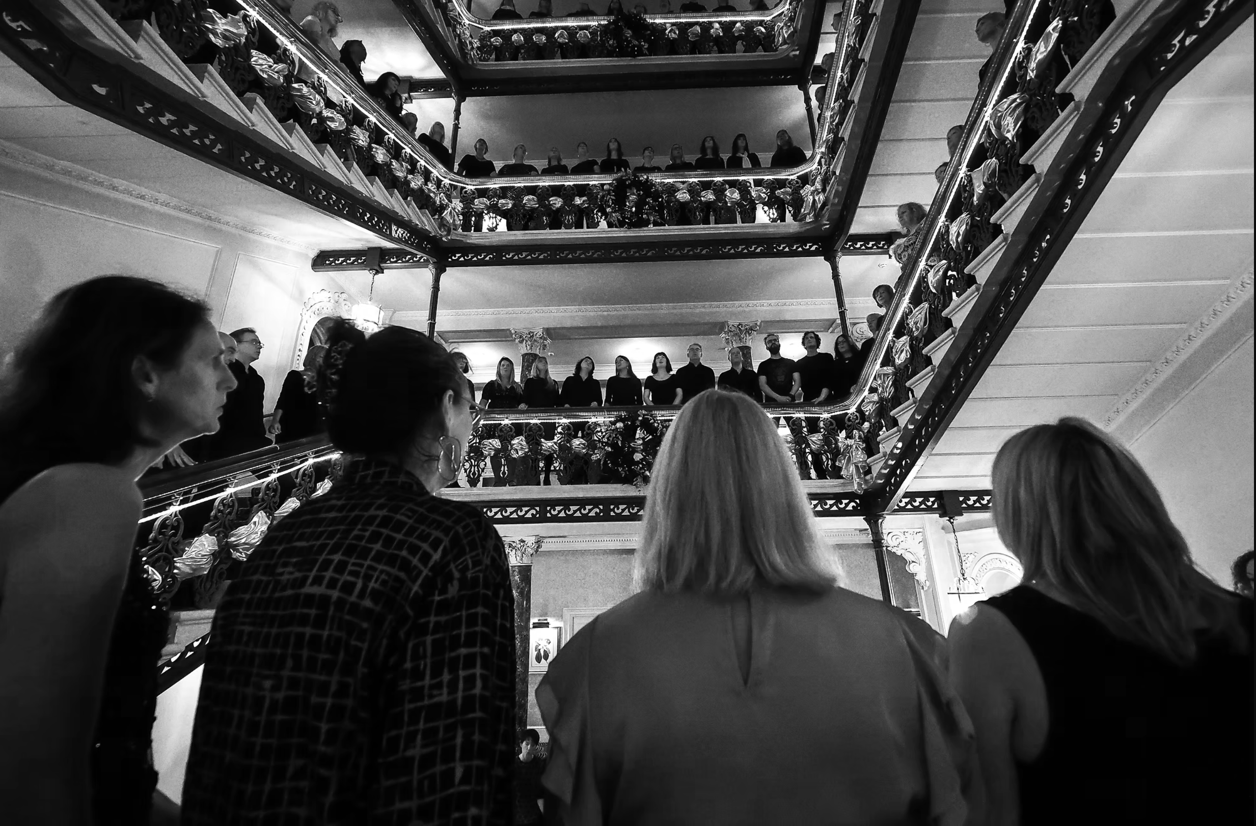 Soul of the City Choir members lining the Grand Hotel stairwell for a joyful flash-mob performance, voices rising through the central space.