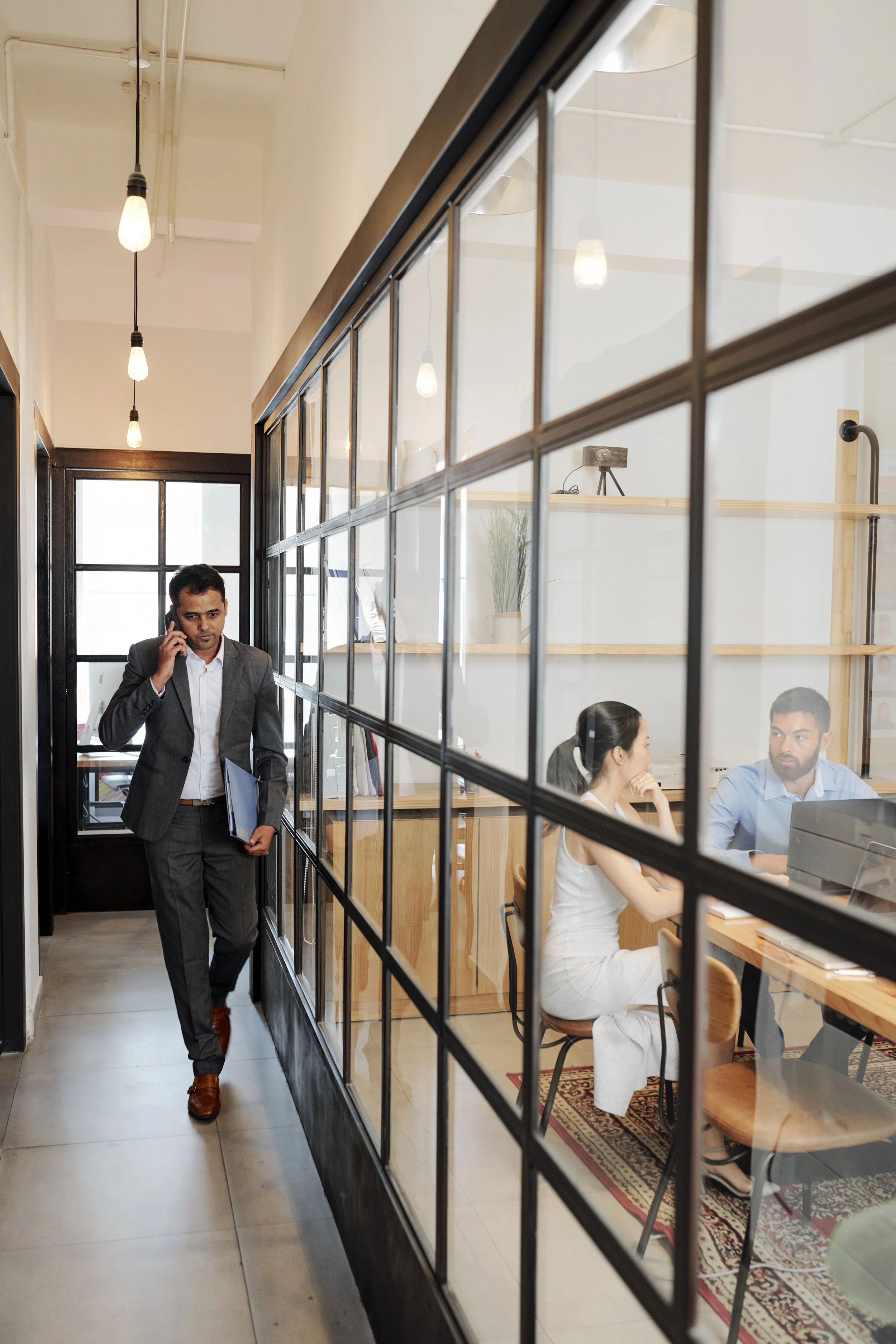 Man in gray suit walking and talking on cellphone in front of a glass partition with a meeting room interior visible behind it where two people are seated at a table.