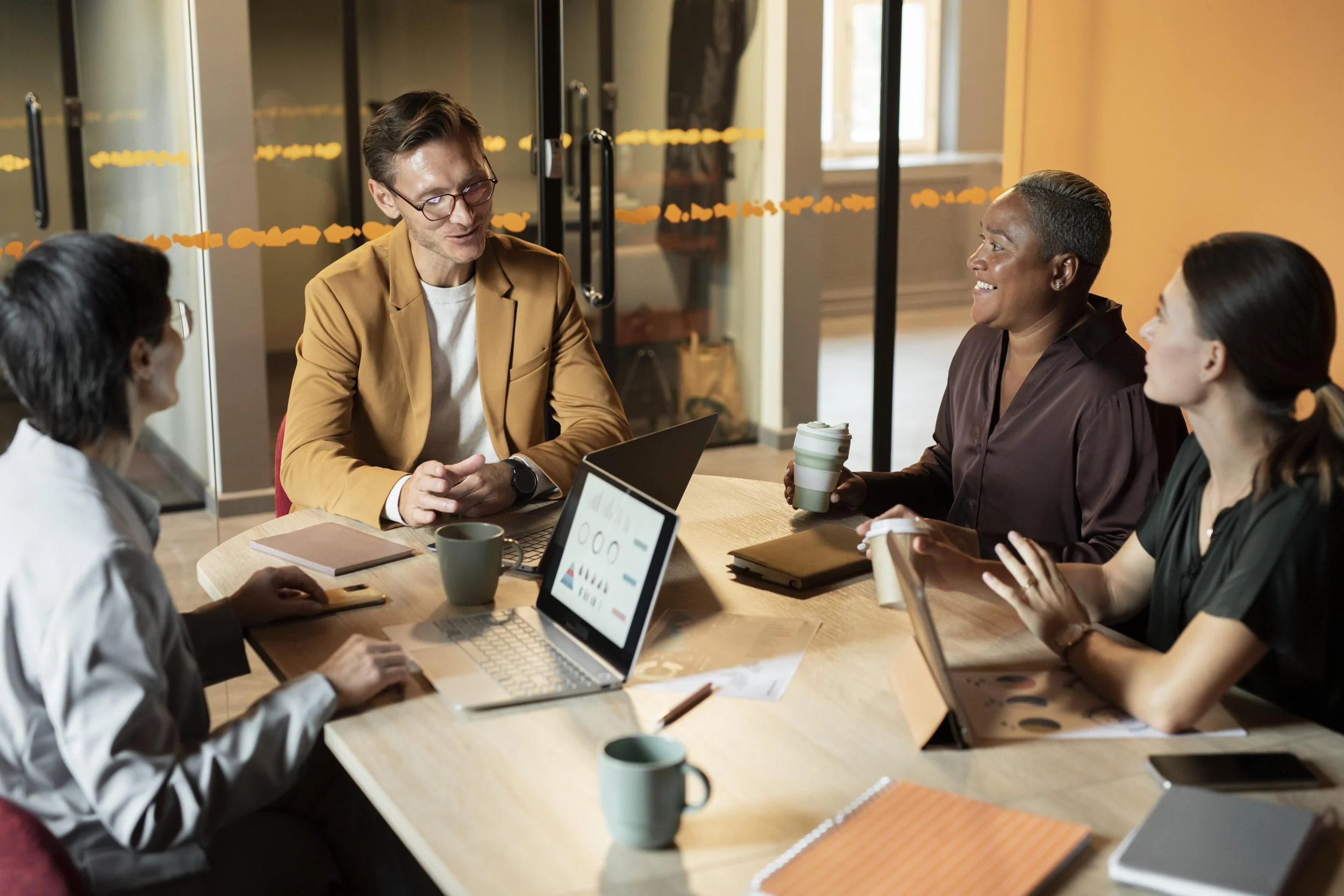 Four diverse professionals having a meeting in a modern office conference room, with laptops, notebooks, and coffee cups on the table.