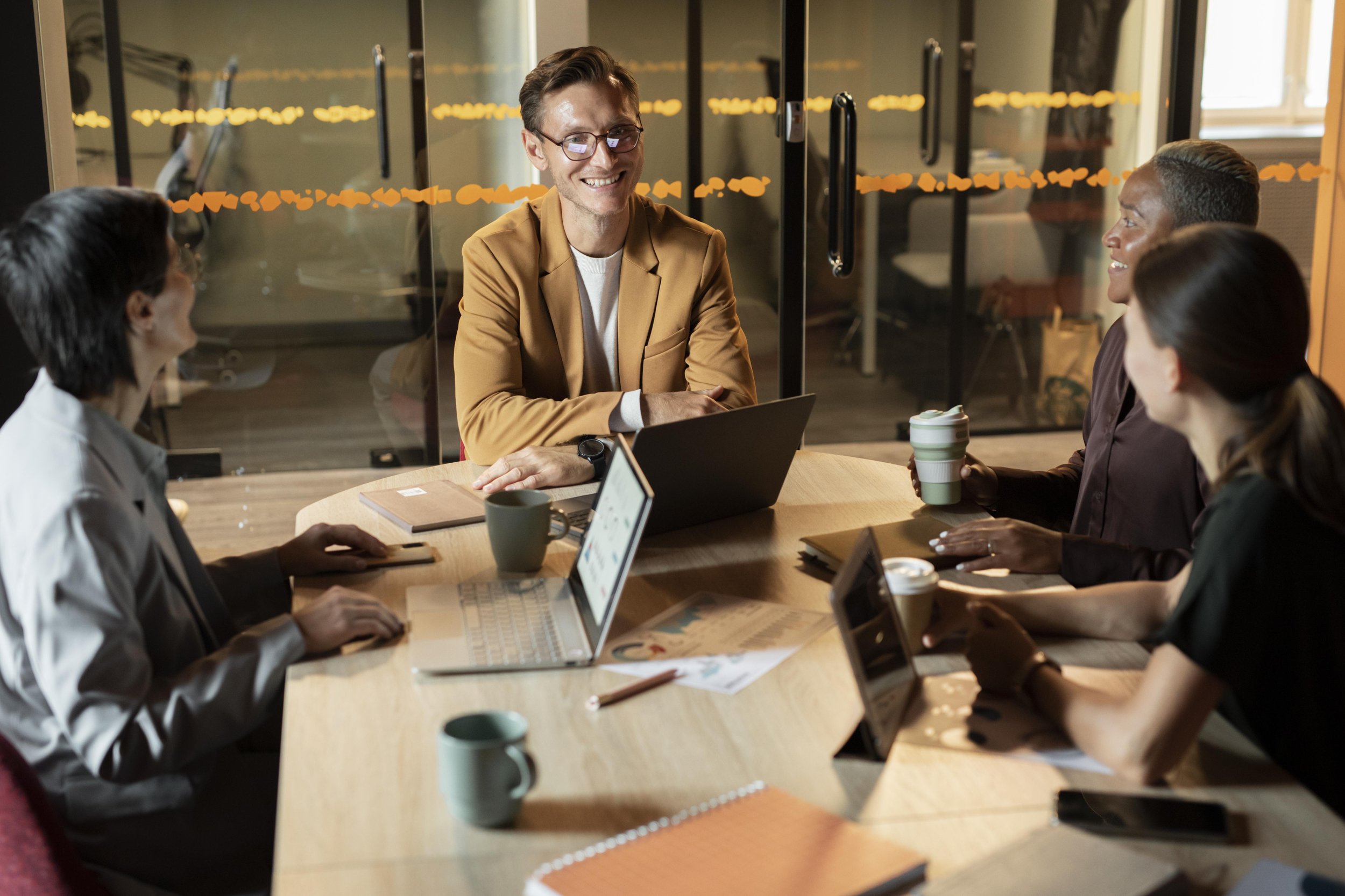 A diverse group of four people having a meeting around a wooden conference table in an office. The man at the head is smiling, wearing glasses and a tan blazer, with a laptop in front of him. Two women and one man are listening; one woman is holding a coffee cup, another woman has a tablet, and the man is sitting with a coffee mug. There are laptops, notebooks, and coffee cups on the table.