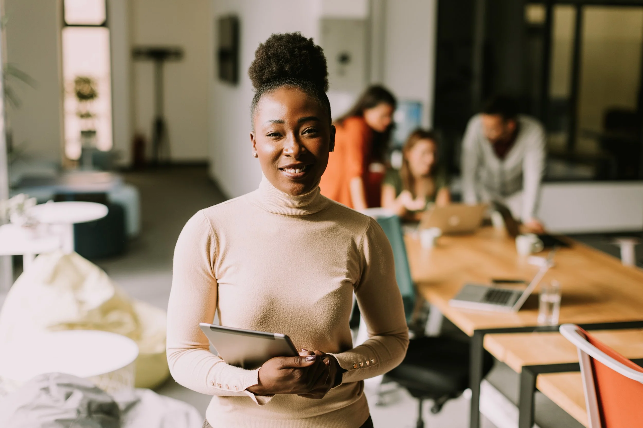 A woman holding a tablet, smiling, in a modern office with others working around a large table.