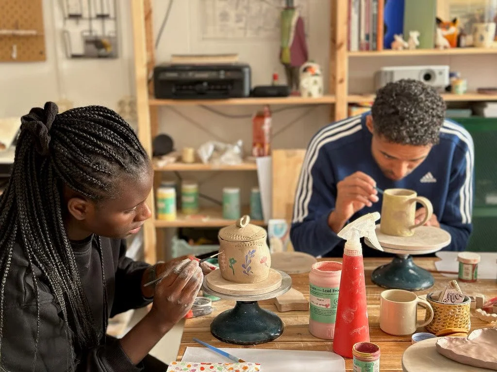 Two people working on clay pottery at a woodworking table in a workshop. The person on the left has braided hair and is painting or carving a pottery piece, while the person on the right is using a tool on a pottery mug.