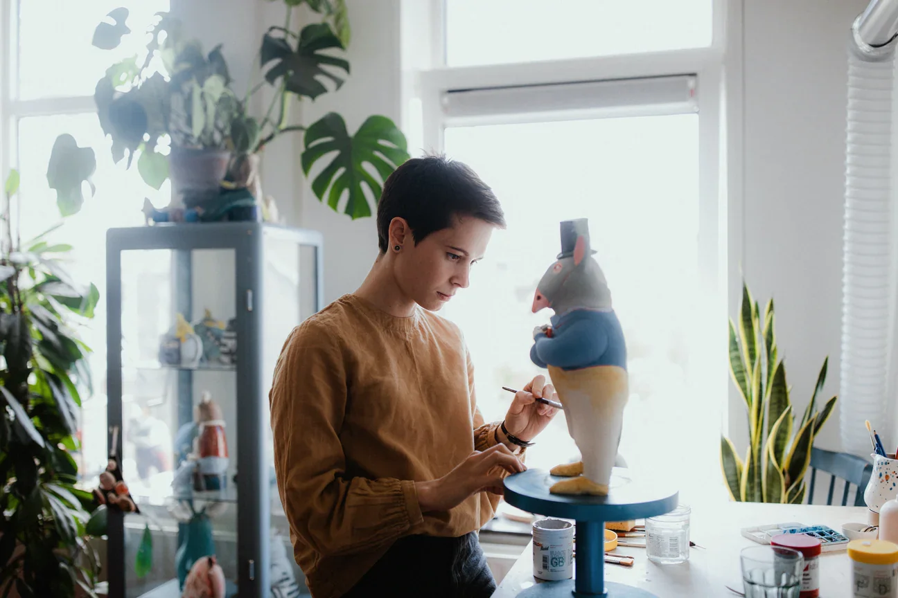 A woman painting a sculpture of a yak in a well-lit art studio filled with plants and art supplies.