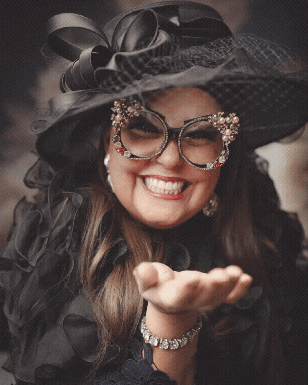 A woman dressed in a black outfit with ruffled details, wearing large decorative glasses adorned with pearls and rhinestones, smiling broadly, and blowing a kiss. She has on large earrings, a pearl bracelet, and a large ornate hat with ribbons and netting.