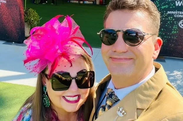 A smiling woman and man wearing sunglasses outdoors, with the woman wearing a bright pink fascinator hat and the man dressed in a tan jacket with a floral tie.