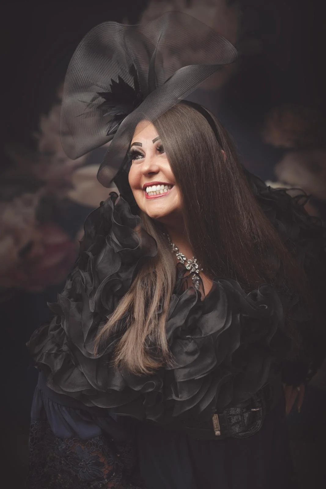 A woman smiling, wearing a large black hat with feathers, a black ruffled dress, and a statement necklace.
