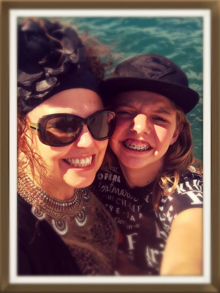 A woman and a young boy smiling and posing for a selfie in front of a body of water. The woman is wearing sunglasses, a black headscarf, and a statement necklace. The boy is wearing a black cap and braces, and has long hair.
