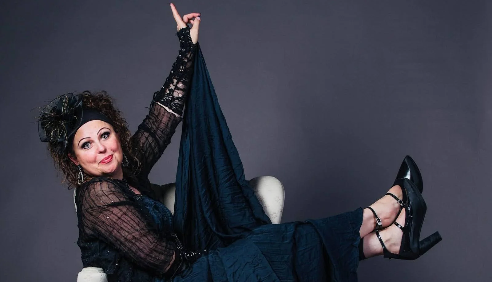 A woman with curly hair and vintage style clothing, sitting on a white chair with her legs up, wearing black high heels, a black headband with a bow, and earrings, against a dark gray background.