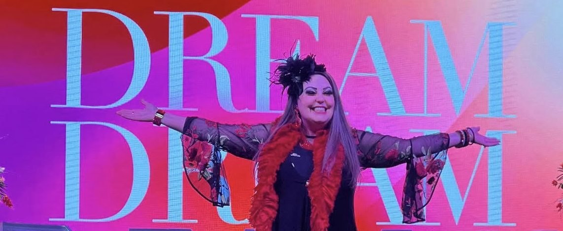 A woman standing in front of a colorful background with the words 'DREAM BELIEVE' and smiling while spreading her arms.