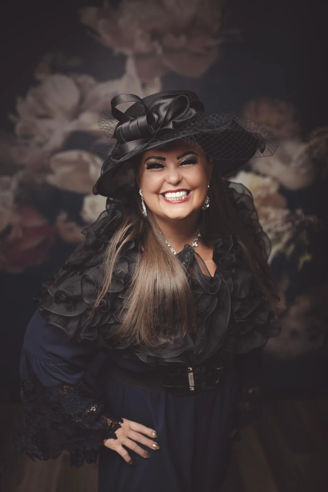Smiling woman wearing a large black hat with lace and bow details, lace dress, and pearl jewelry in front of a floral backdrop.