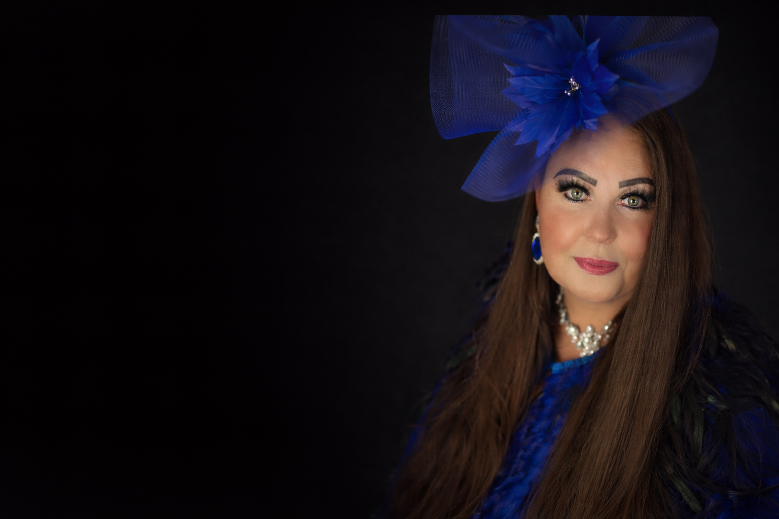 connie mannering Portrait of a woman with long brown hair, wearing a large blue fascinator, earrings, and a necklace, against a black background.