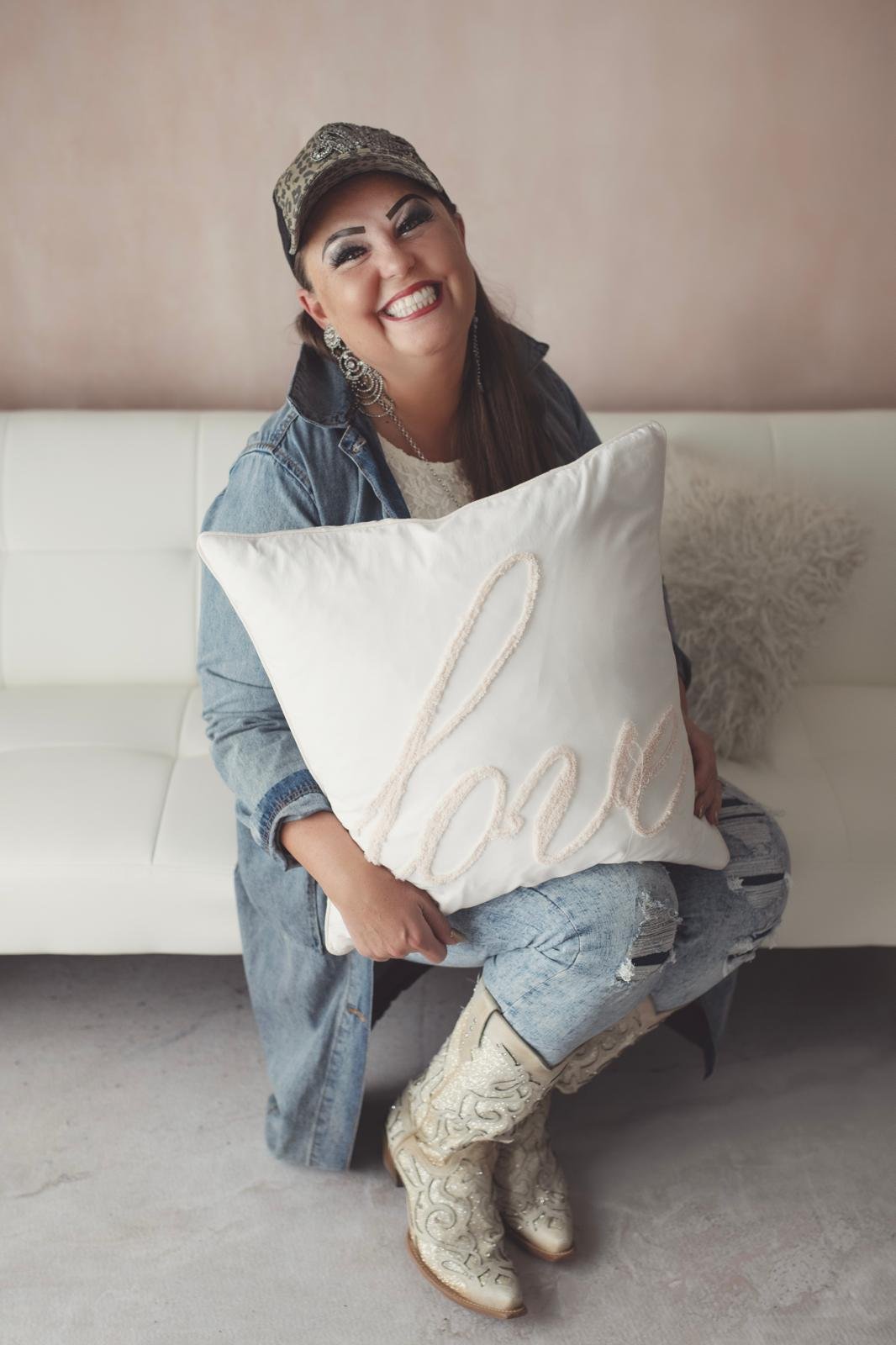 Woman smiling and sitting on a white couch, holding a large white pillow with the word 'love' written on it, wearing cowboy boots, ripped jeans, a denim jacket, and a cap.