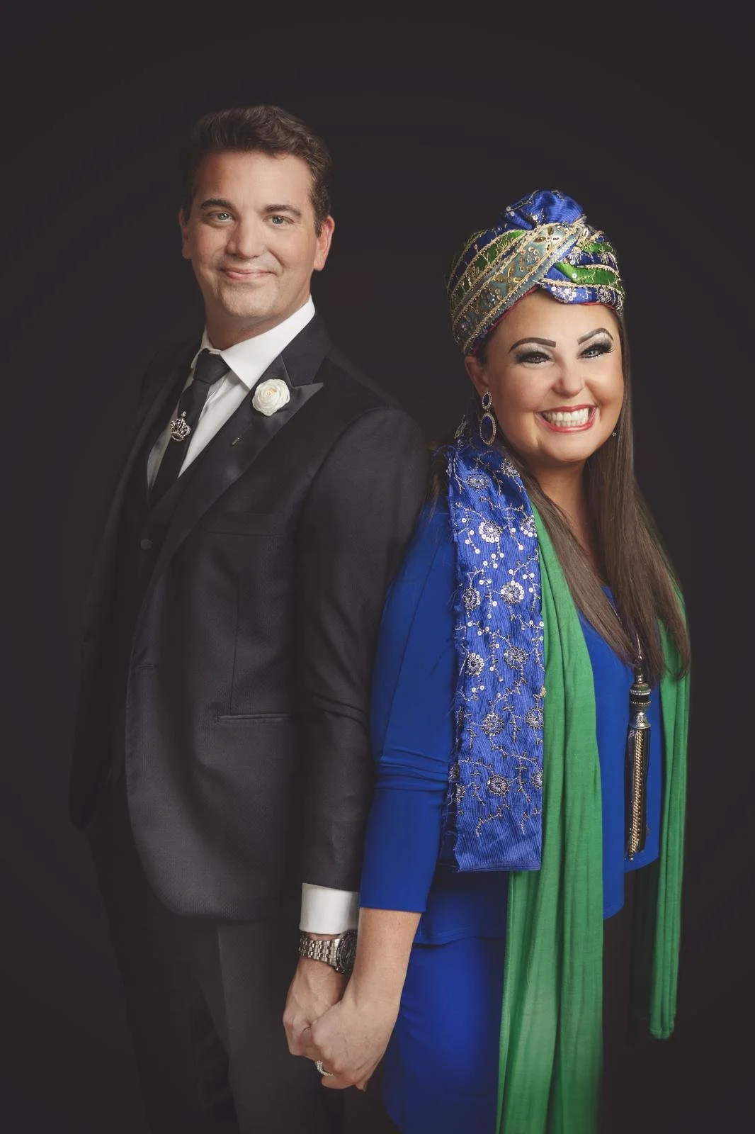 A man in a black suit and a woman in a colorful traditional outfit, standing back-to-back, smiling, against a black background.