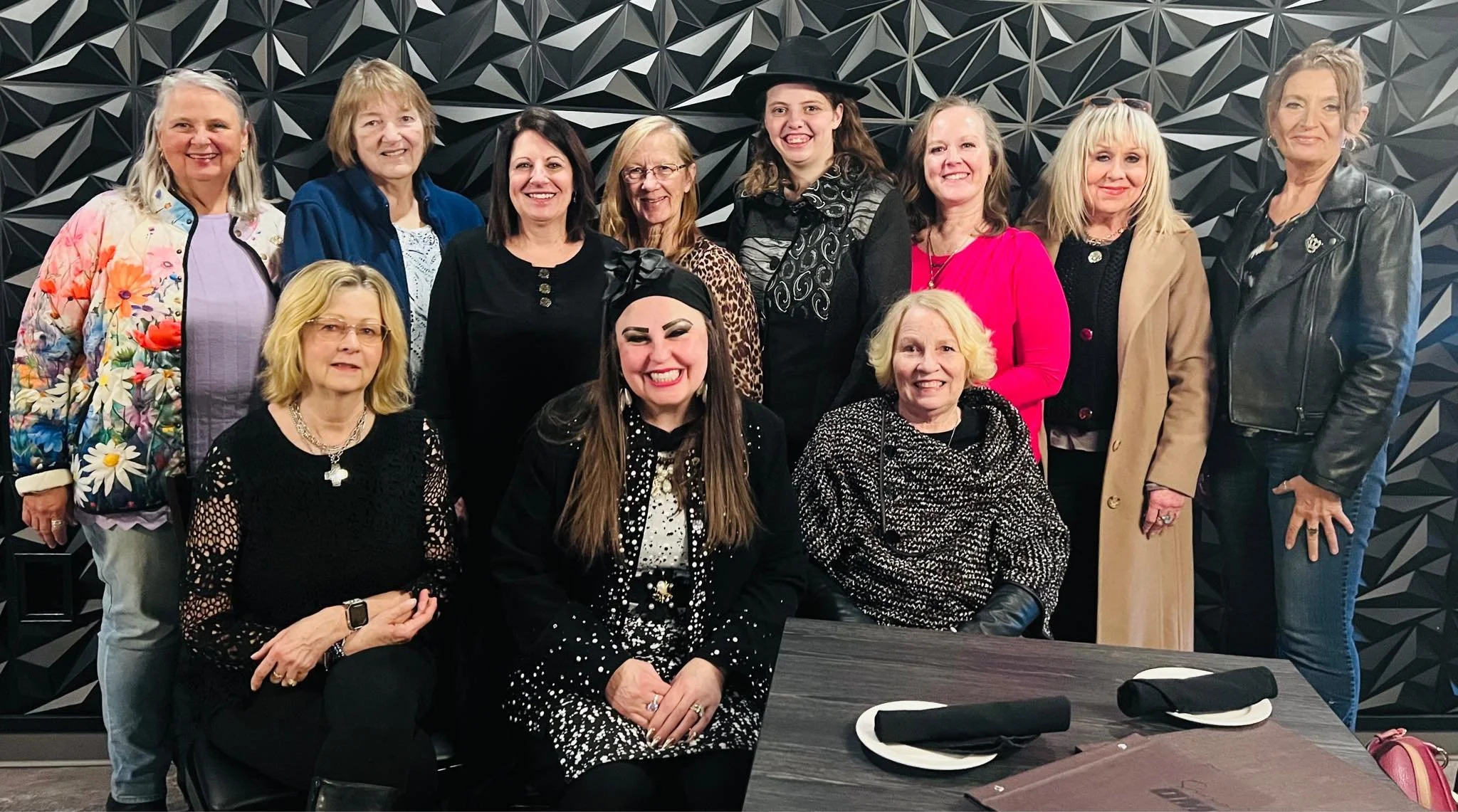 A group of twelve women, some sitting and some standing, pose together in a restaurant or event space with a black geometric patterned wall behind them. They are smiling and dressed in colorful and fashionable attire.