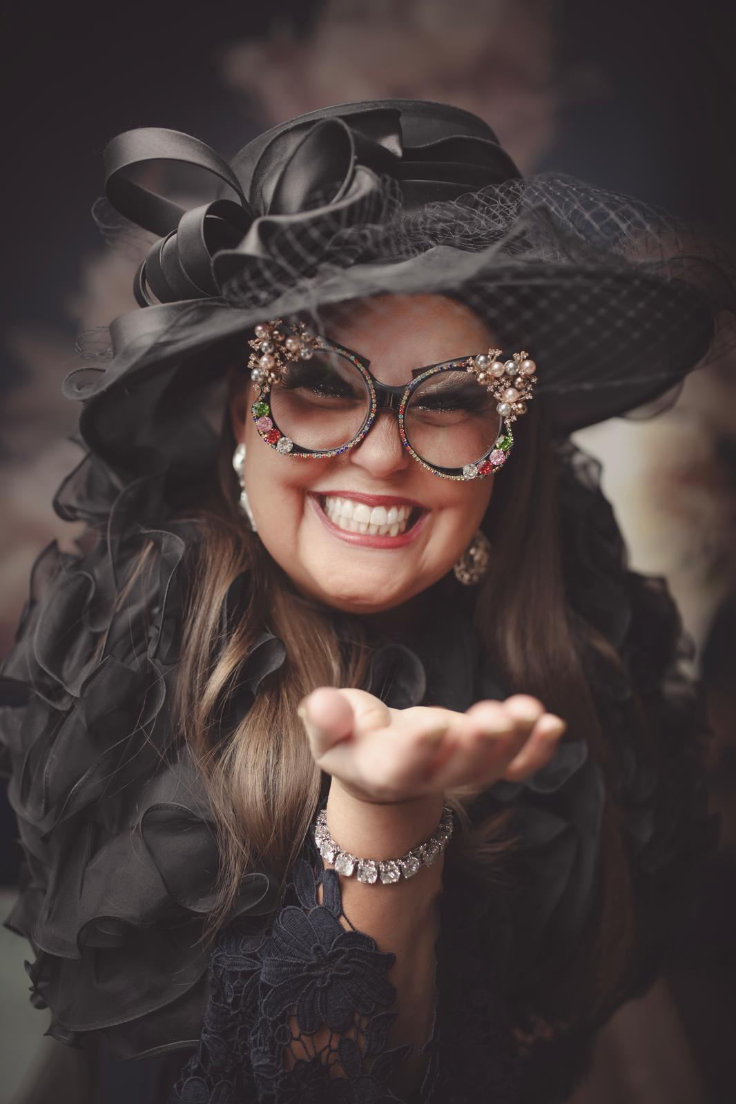 Happy woman in fancy black hat and glasses, smiling and blowing a kiss.