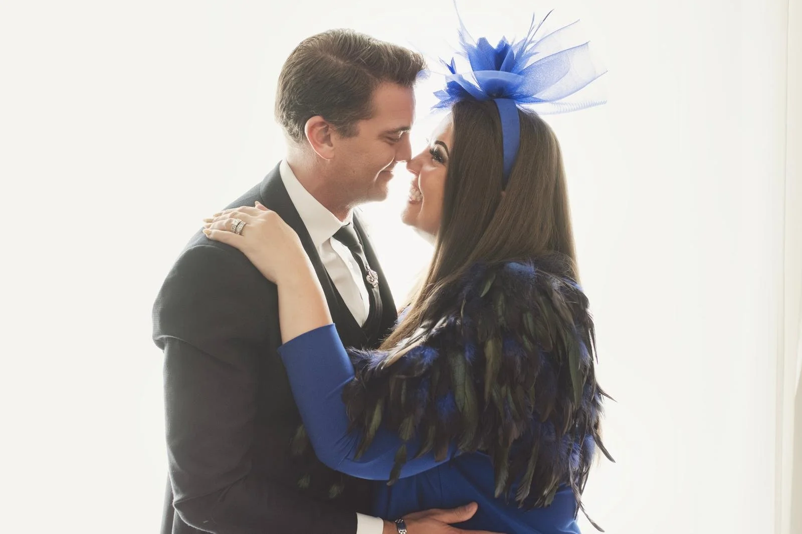 A man and woman are smiling closely at each other, with their foreheads touching. The man is wearing a black suit, and the woman is dressed in a blue outfit with feather details and a matching blue headband with a decorative tulle accessory. The back
