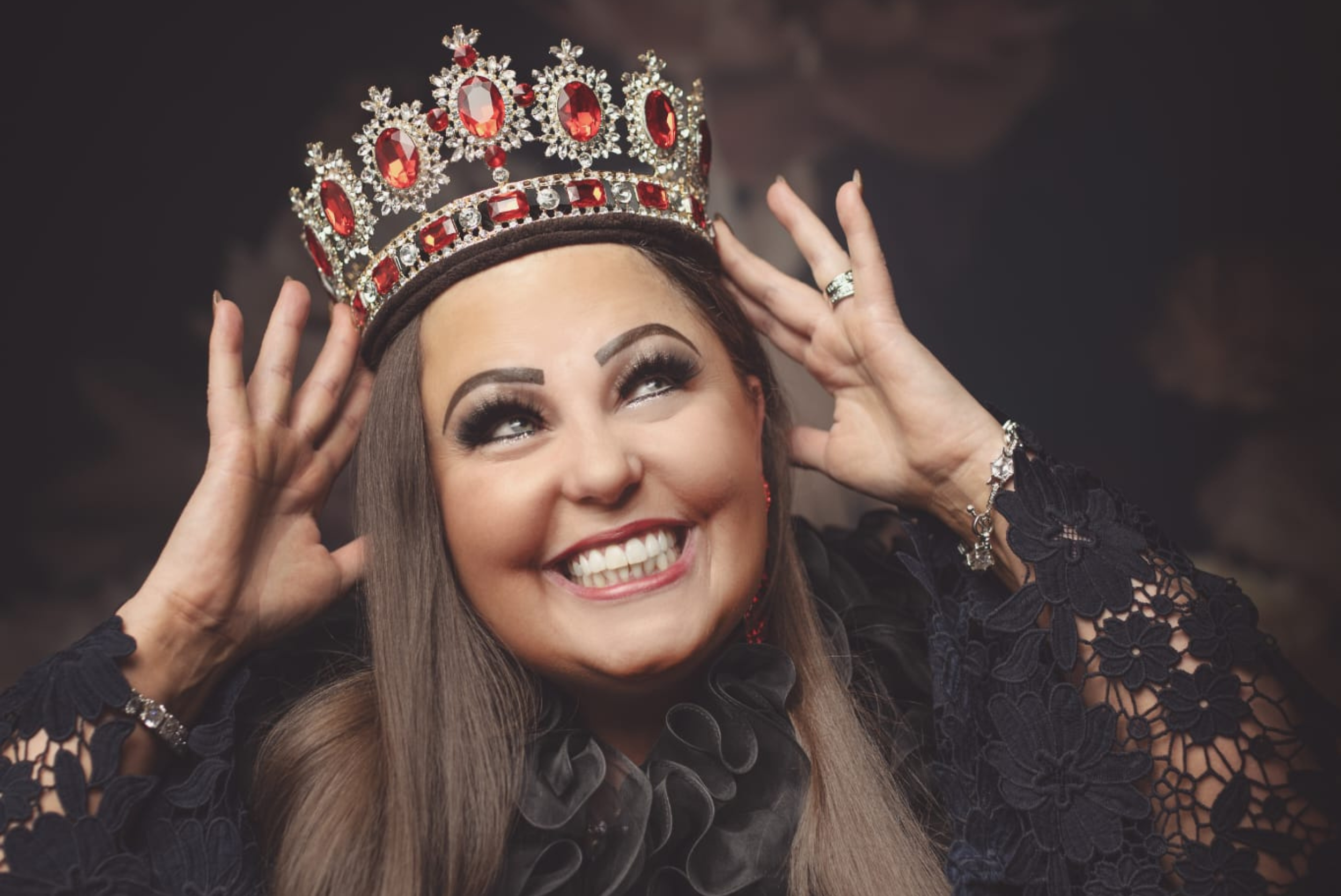A woman with long brown hair wearing a black lace dress and a sparkling bracelet, smiling widely and holding a large crown decorated with red and clear gemstones on her head.