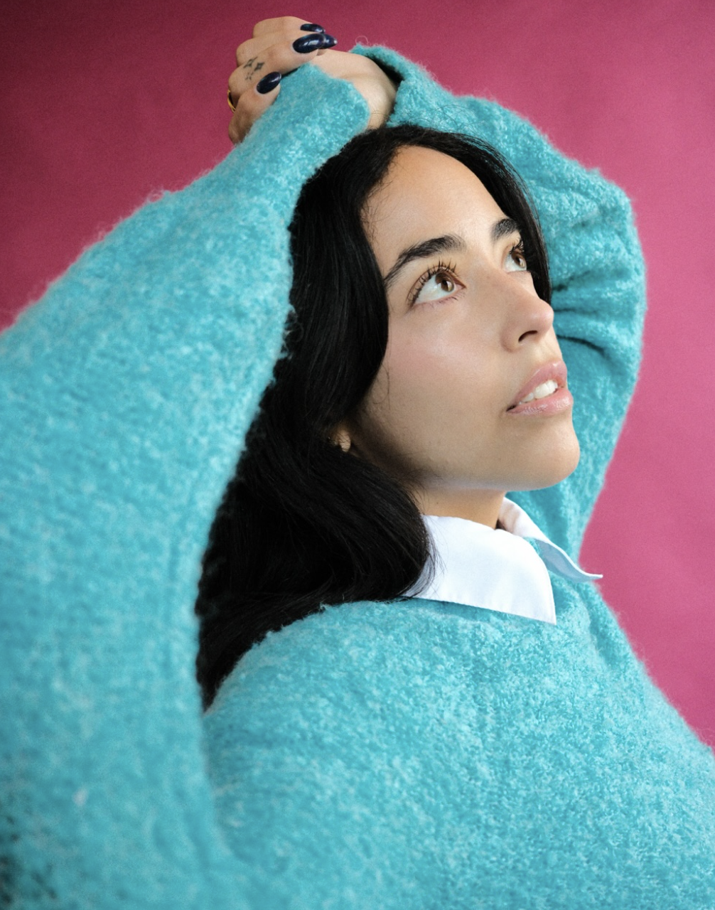 A woman with dark hair and light skin, wearing a turquoise fuzzy sweater, looking upwards against a pink background.