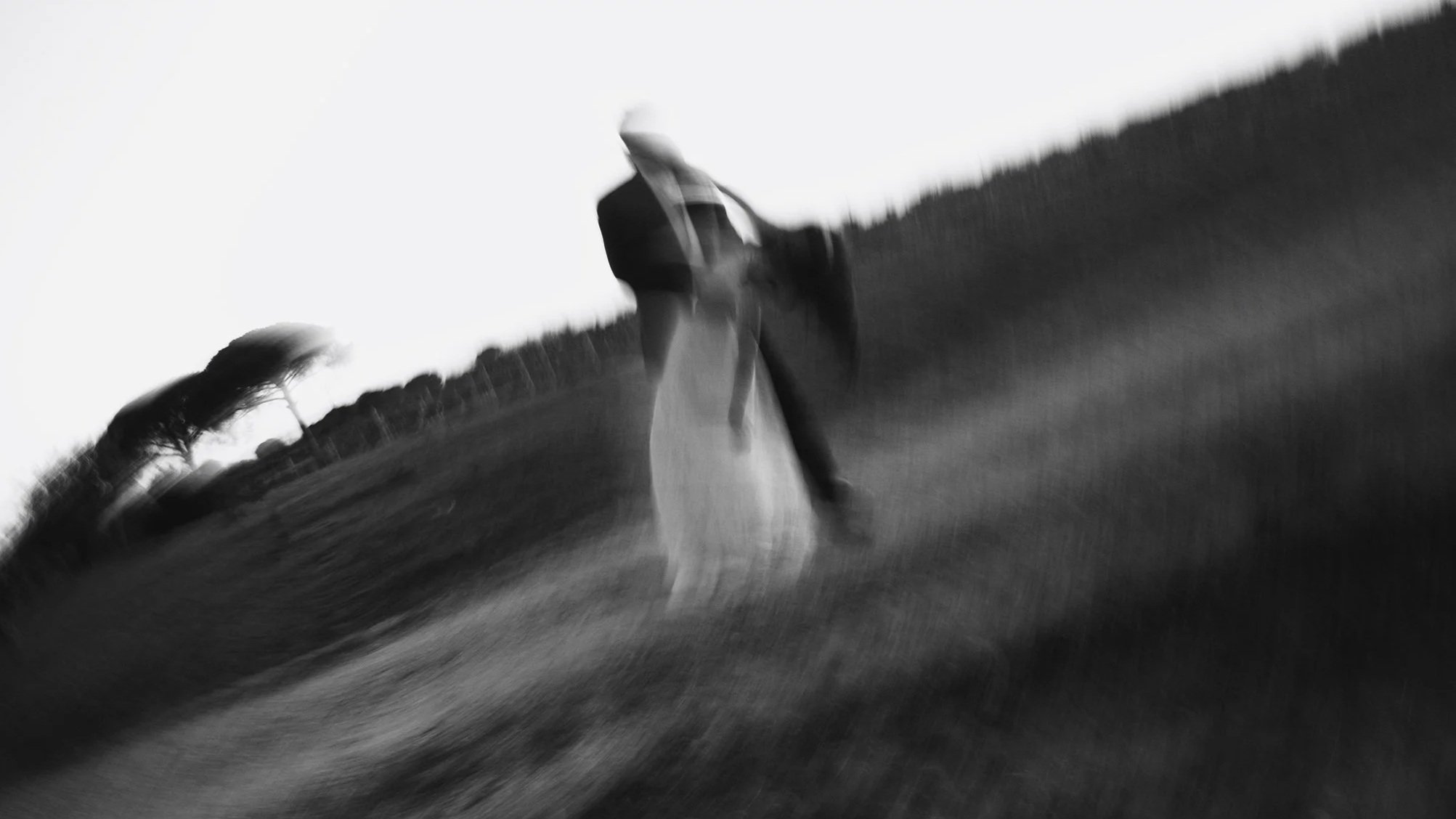 A blurred black-and-white photo of a person in formal attire standing on a grassy hill, holding a long flowing veil or fabric with trees in the background.