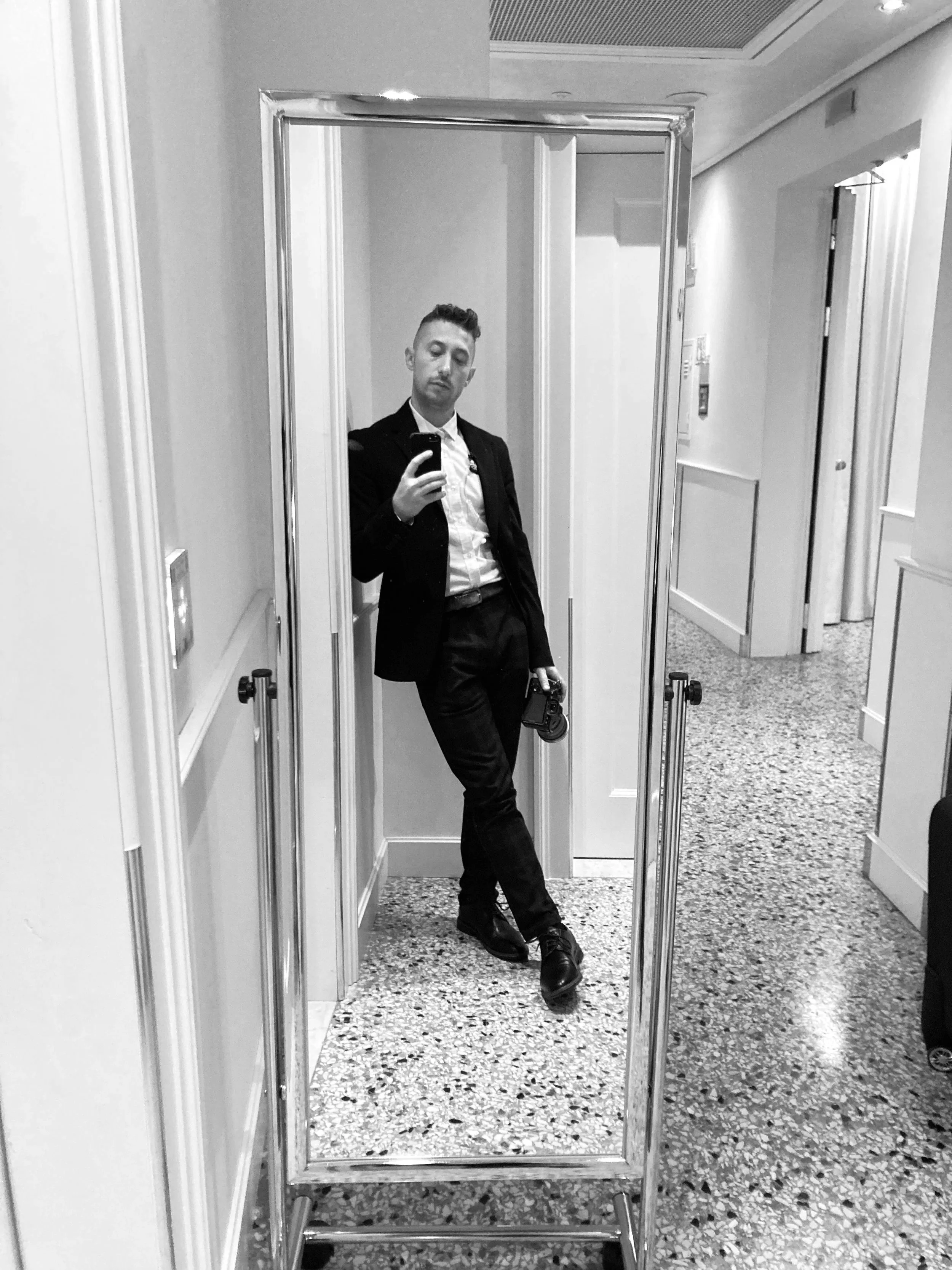 A man taking a mirror selfie in a hotel hallway, dressed in a dark suit and white shirt, holding a camera, with a neutral expression.