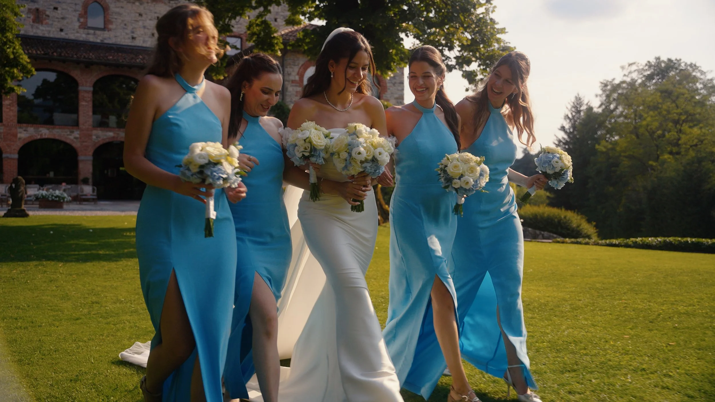 A bride and her four bridesmaids walk outdoors on a sunny day, holding bouquets and smiling.