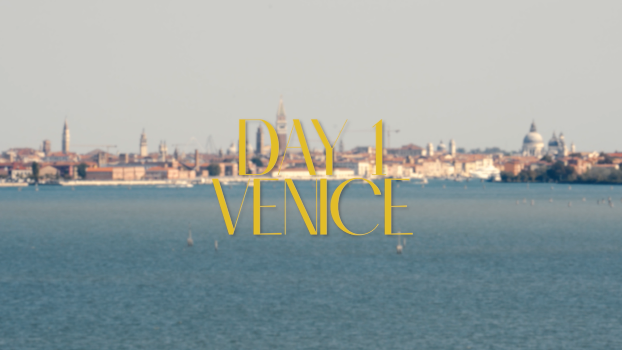 Sunset over the Venice skyline with iconic buildings and water in foreground, overlaid with the text 'Day 1 Venice' in yellow.