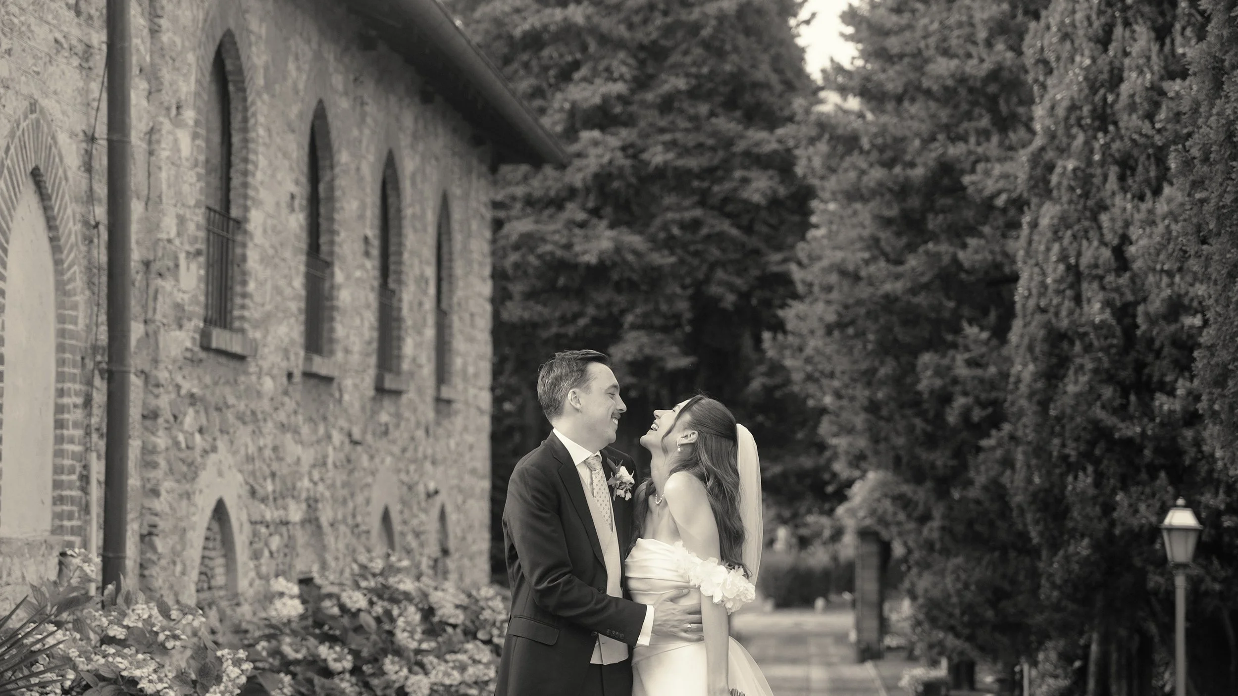 A black-and-white photo of a bride and groom smiling and looking at each other outdoors. The groom is in a suit, and the bride is in a wedding dress with a veil, standing in front of a stone building with arched windows and surrounded by trees and flowers.