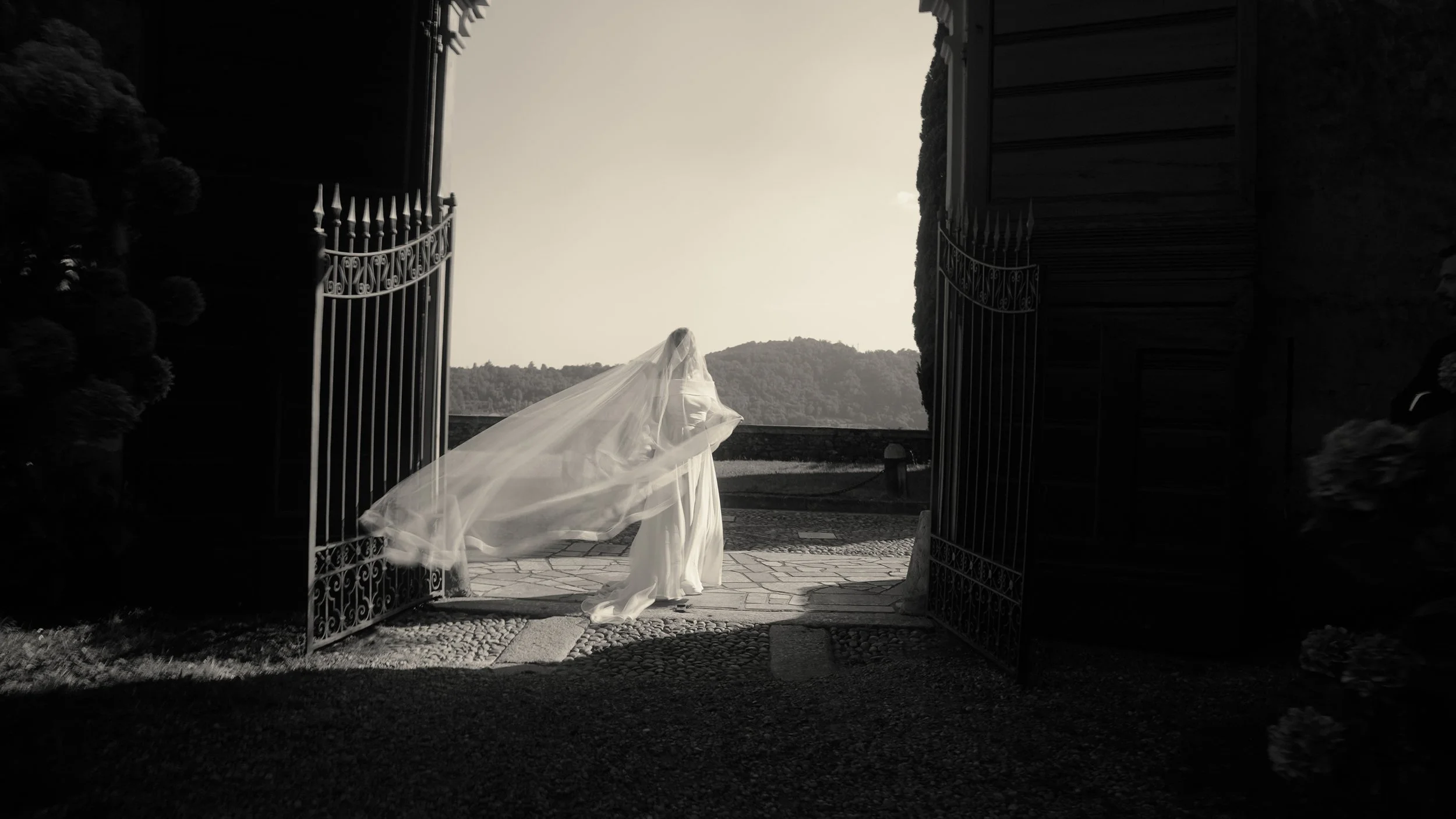 Bride in wedding dress with veil walking through open gates onto scenic outdoor area.