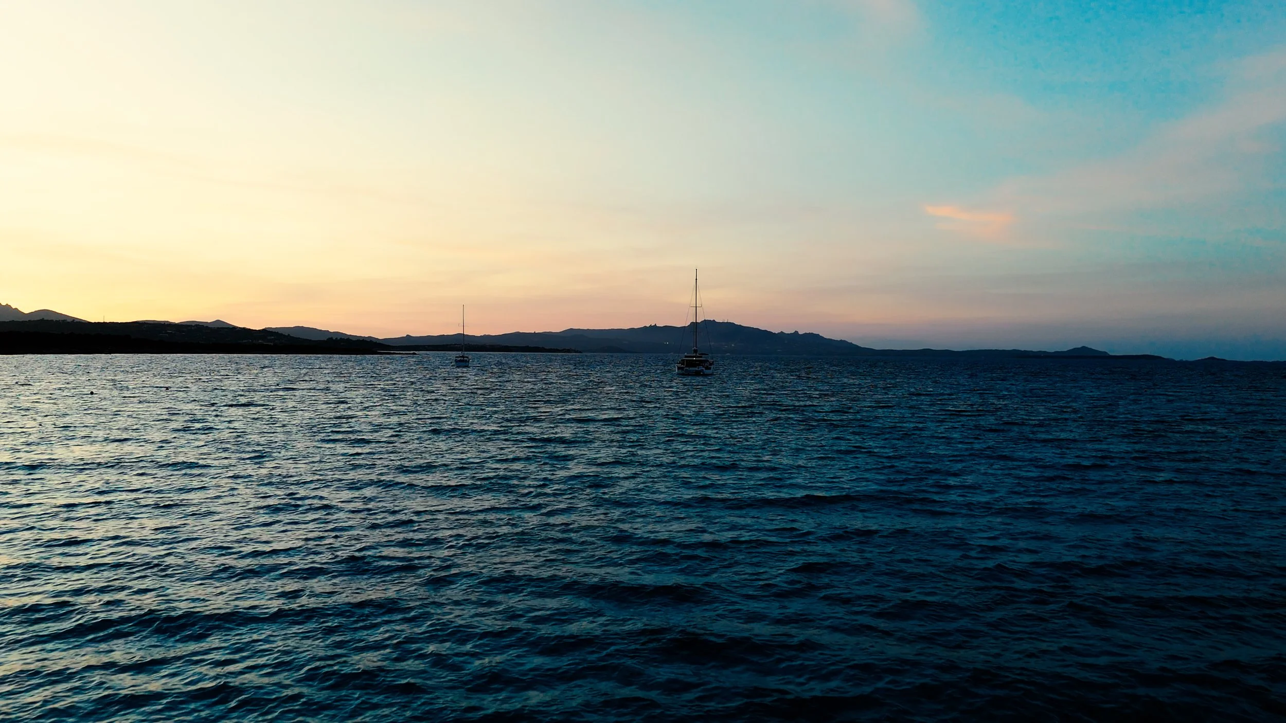 Calm ocean water with two sailboats and a mountainous coastline at sunset or sunrise with soft pastel sky.