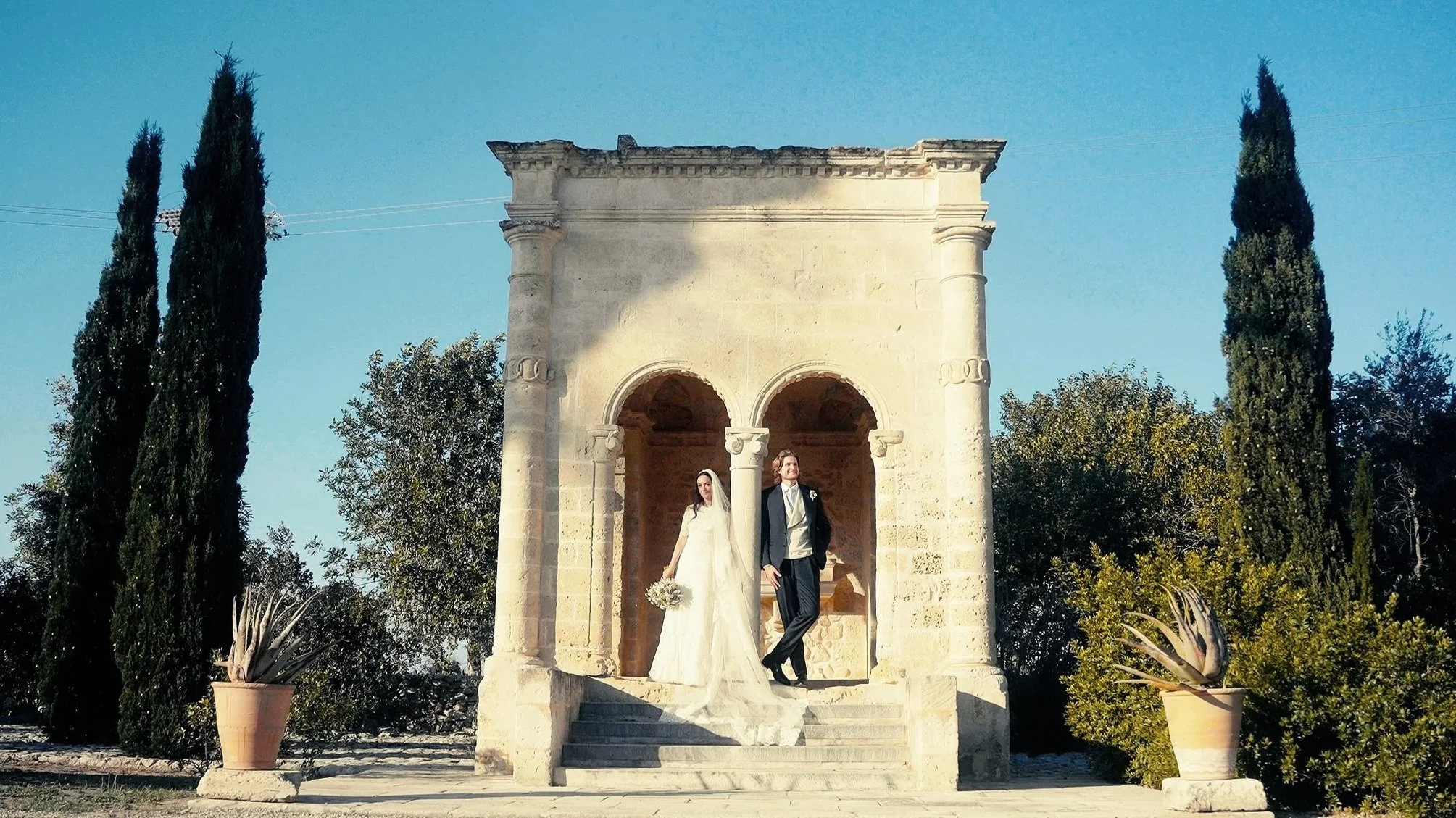 Bride and groom standing on steps of a stone pavilion, with tall cypress trees on either side, under a clear blue sky.