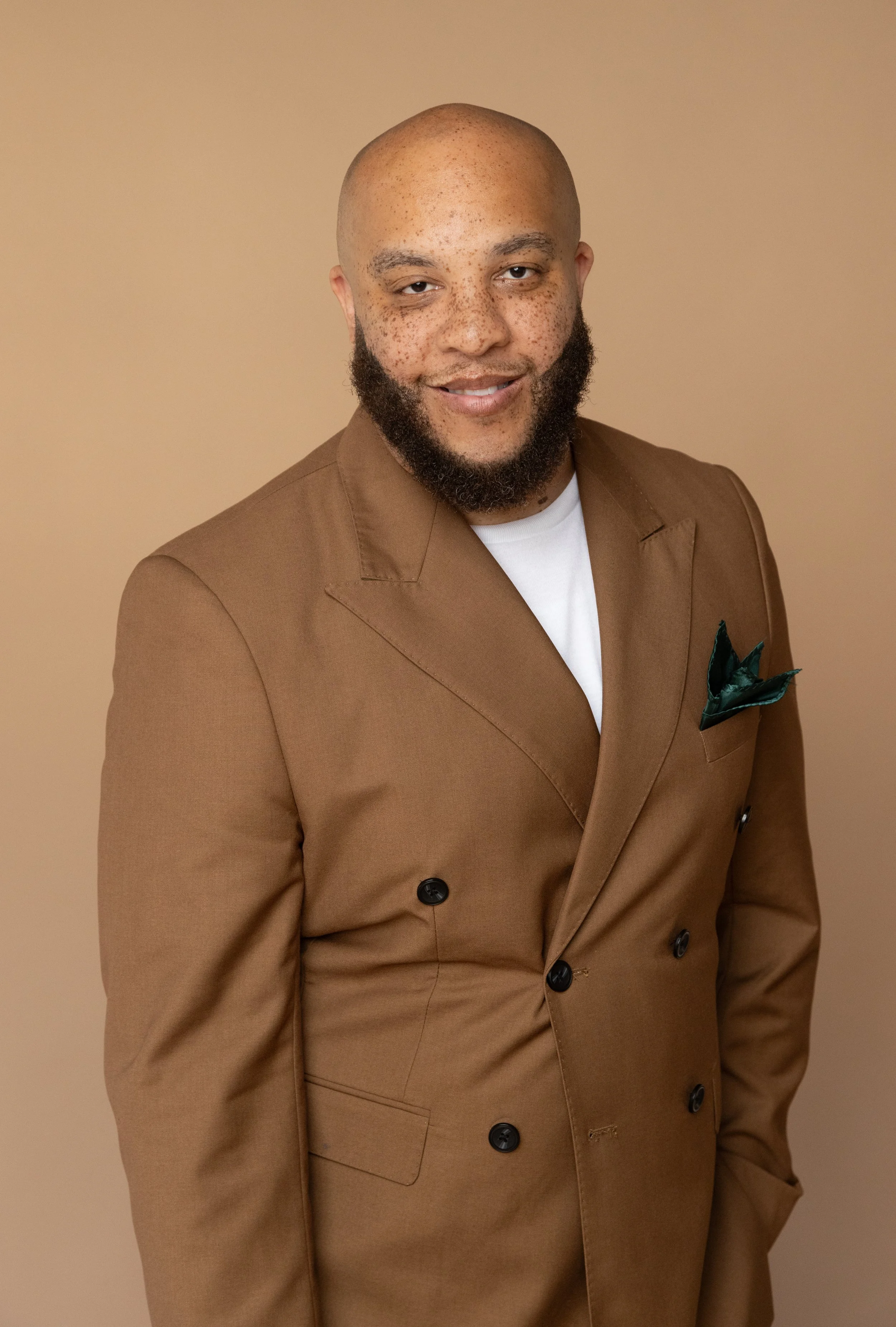 A man with a shaved head and beard wearing a brown double-breasted suit jacket over a white shirt, standing against a beige background.