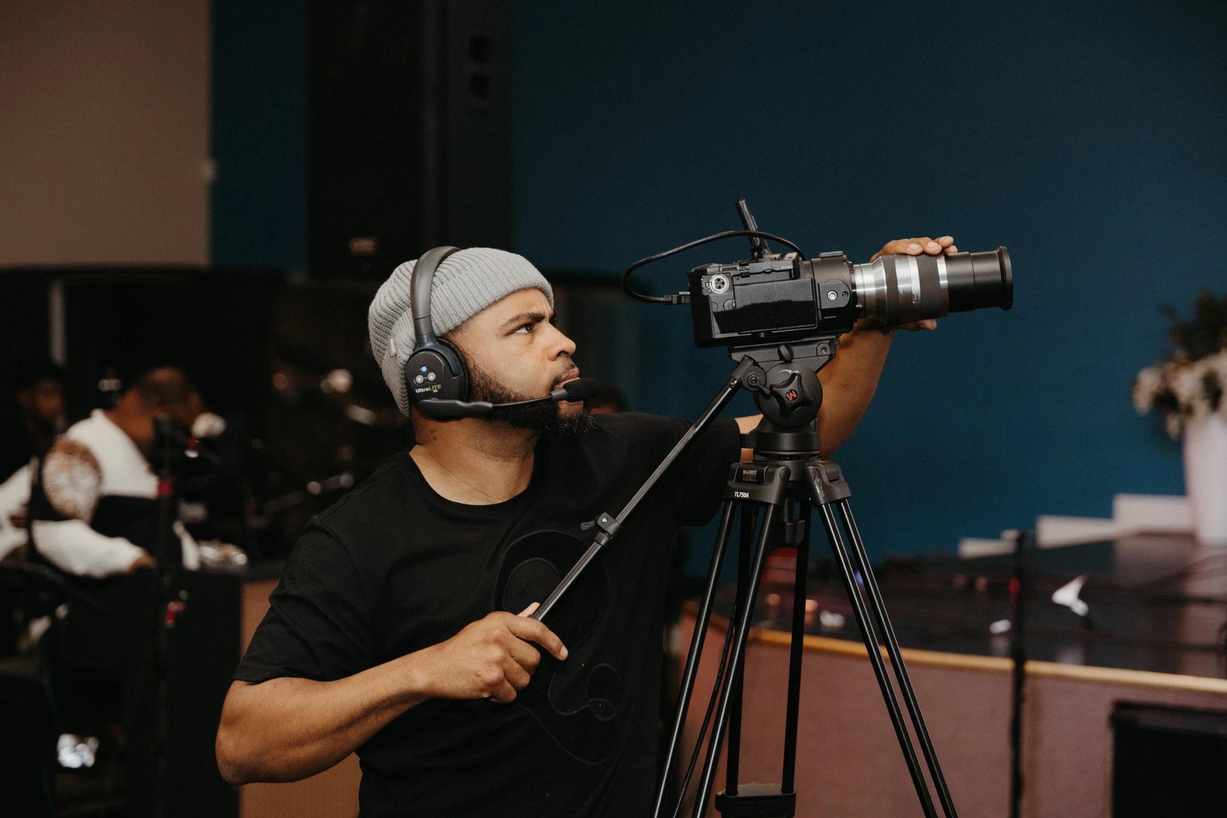 A man wearing a gray beanie and black shirt operates a professional video camera on a tripod with headphones on, filming in an indoor setting with a blue wall in the background.