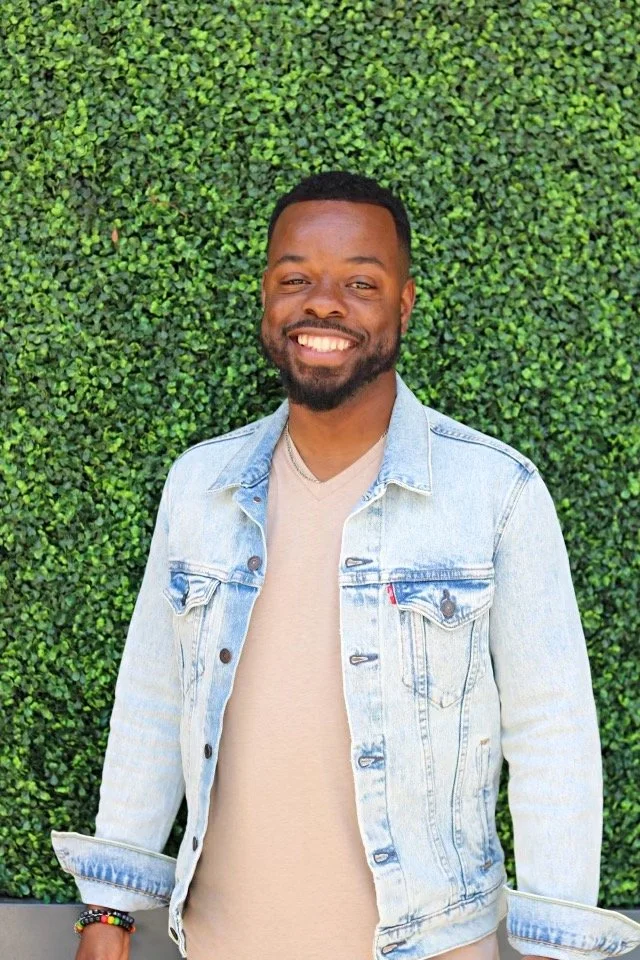 A smiling man standing in front of a lush green hedge, wearing a light denim jacket over a beige shirt, with a bracelet on his left wrist.